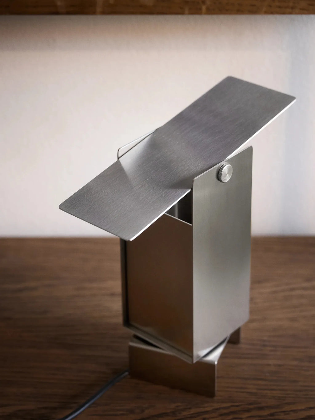 MOEBE Pivoting Table Lamp, a sleek modern minimalist desk lamp crafted from brushed stainless steel with a matte silver finish and a unique adjustable geometric design. This industrial chic table lamp features a rectangular base and a pivoting metal shade, allowing for directional light, perfect for contemporary home offices, Scandinavian living rooms, or minimalist bedside tables.