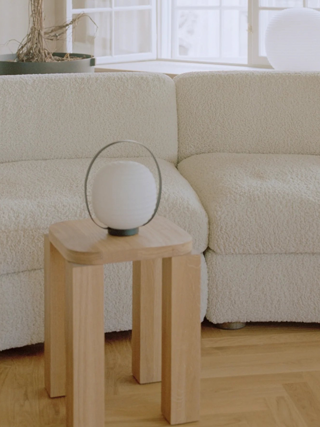 New Works Lantern Portable Lamp, a premium white frosted glass globe with radiating texture, supported by a dark wire handle & base on a light solid wood stool, in a modern interior. Ideal for ambient lighting.