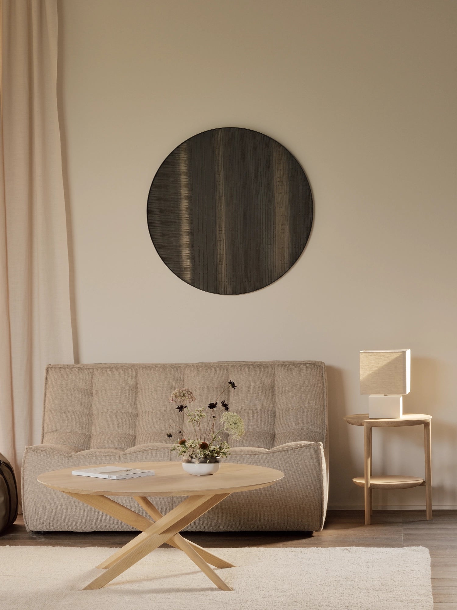 Ethnicraft Mikado Coffee Table, a round, light oak table with a distinctive criss-cross leg design, placed on a light rug in a cozy living room setting with a beige sofa.