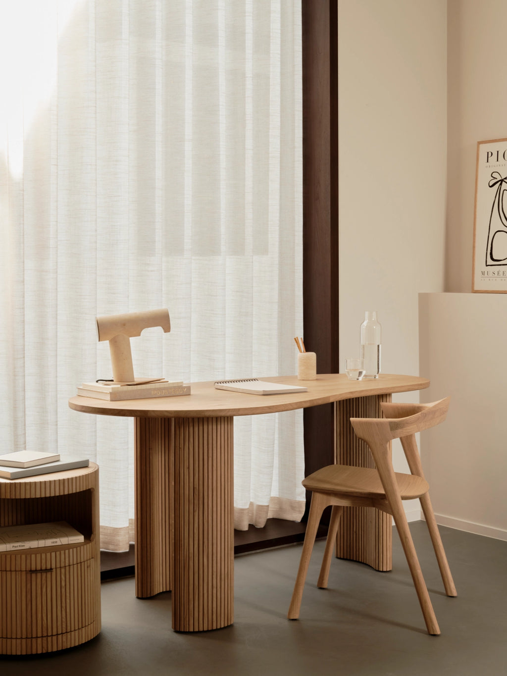 Ethnicraft Boomerang Desk in natural oak, with a light wood oval tabletop and fluted cylindrical legs, paired with a matching oak chair and a round side table in a sunlit room.