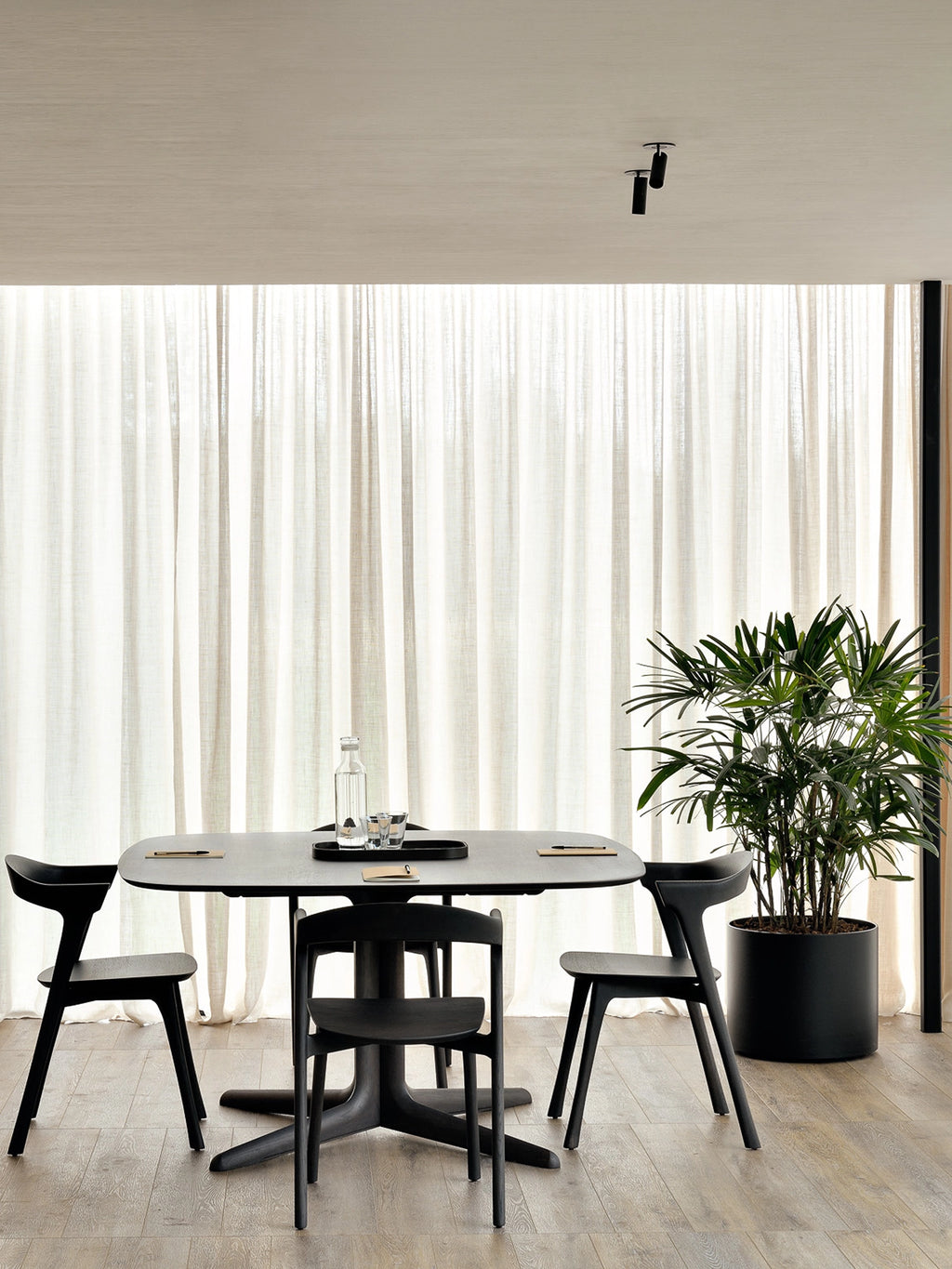 Ethnicraft Corto Dining Table in dark brown oak, square shape, set in a modern dining room with black chairs, a glass bottle, and a plant, against a backdrop of sheer white curtains.
