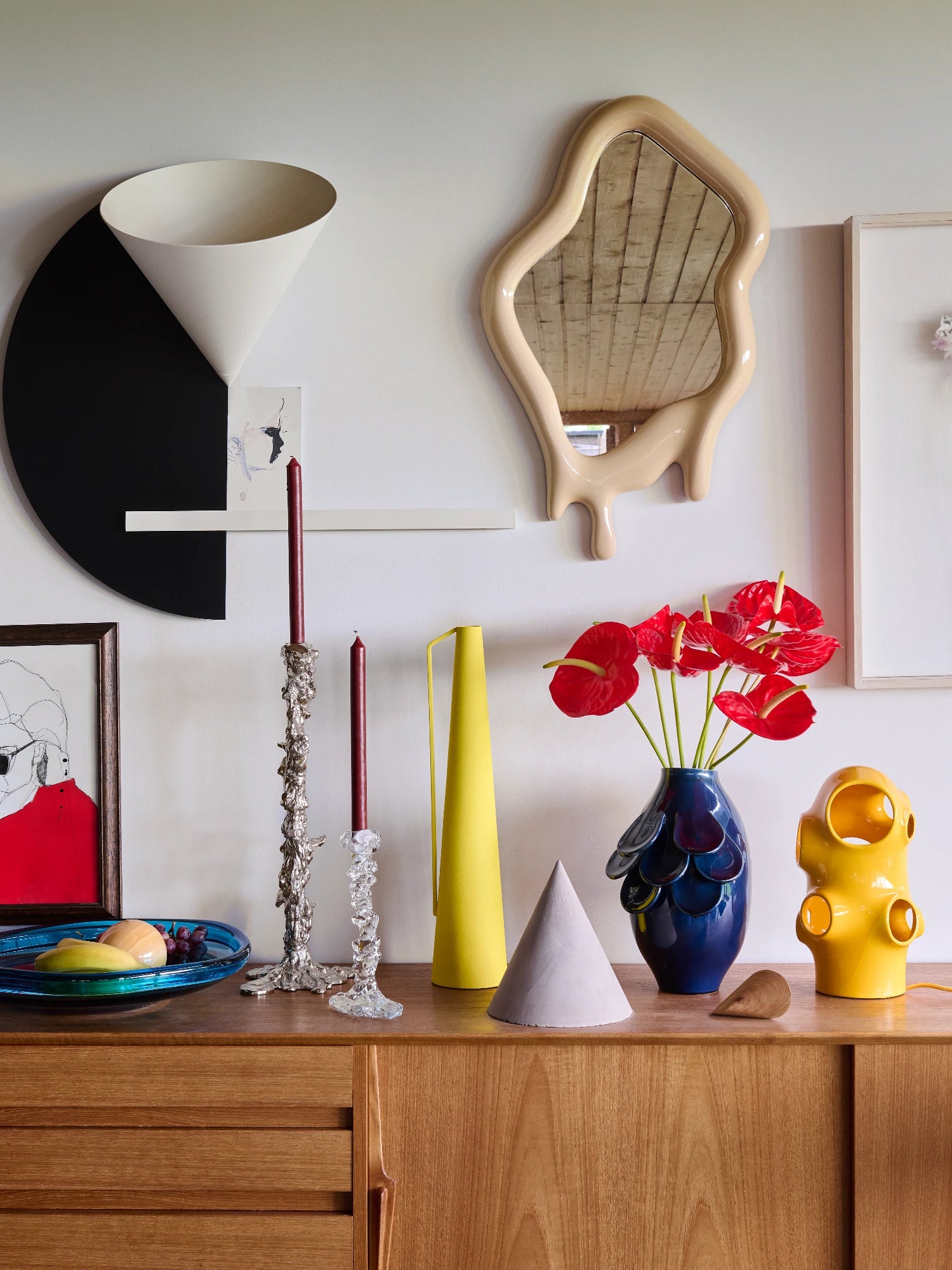 A light beige mirror with a wavy, irregular shape is hanging on a white wall. The bottom of the mirror frame is designed to look like a thick liquid dripping. It's positioned above a wooden sideboard with various art objects, including a blue vase with red anthuriums, two candlestick holders, and a yellow pitcher.