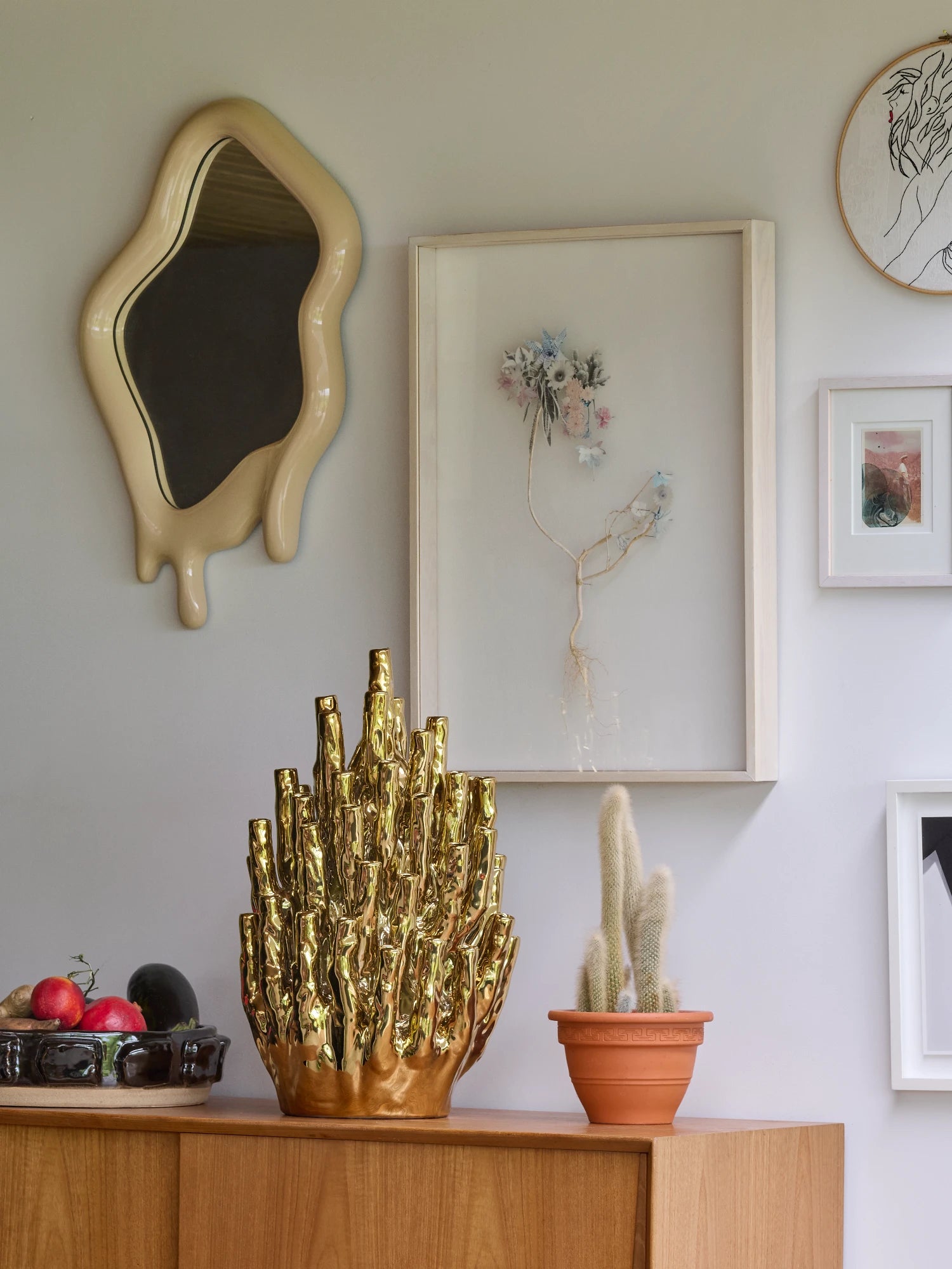 A collection of decorative objects and framed art pieces are displayed on a wooden sideboard and a light-colored wall. A gold, irregularly shaped sculpture is on the sideboard next to a bowl of fruit and a small potted cactus. A unique, beige mirror with a dripping frame is hanging on the wall. Next to the mirror, there is a large framed art print of a plant and other small framed pieces.