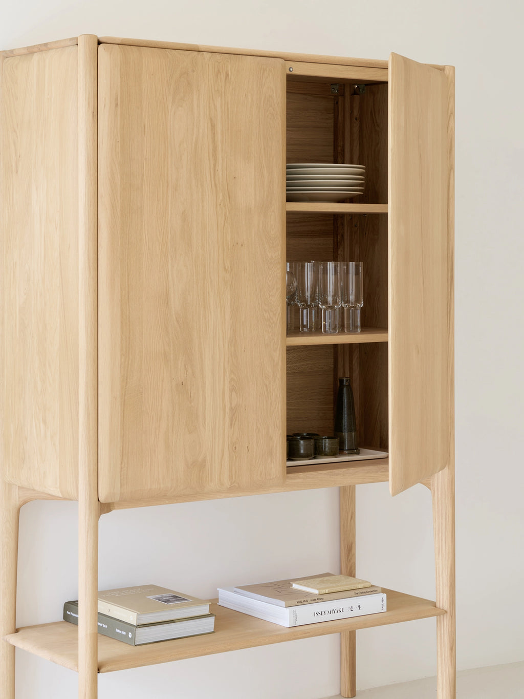 Ethnicraft PI Cupboard, a modern minimalist solid oak cabinet featuring a light natural wood finish with distinctive rounded corners and an elegant vertical grain. This Scandinavian-inspired storage unit includes a closed cupboard section with interior shelves and an open lower shelf supported by slender legs. Perfect as a versatile oak storage cabinet for a contemporary living room, dining room, or entryway.