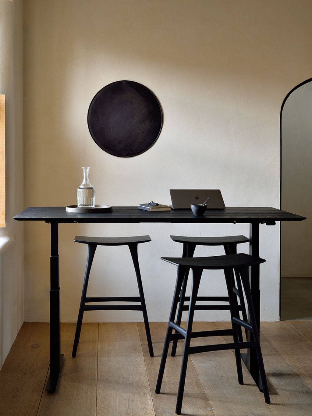 Ethnicraft Bok Adjustable Desk in a rich black finish, featuring a dark tabletop and black adjustable frame, complemented by two black wooden bar stools in a stylish, modern interior.