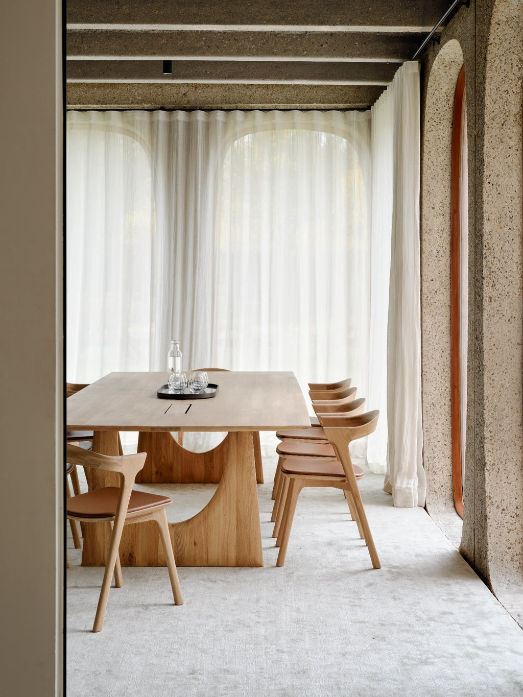 Ethnicraft Geometric Meeting Table, a modern solid oak dining table featuring a sleek rectangular top and a distinctive sculptural base with organic, curved cut-outs. This light natural oak dining table is complemented by matching ergonomic dining chairs with warm tan leather seats, embodying an organic modern aesthetic. Perfect for a contemporary dining room, stylish meeting space, or minimalist interior design.