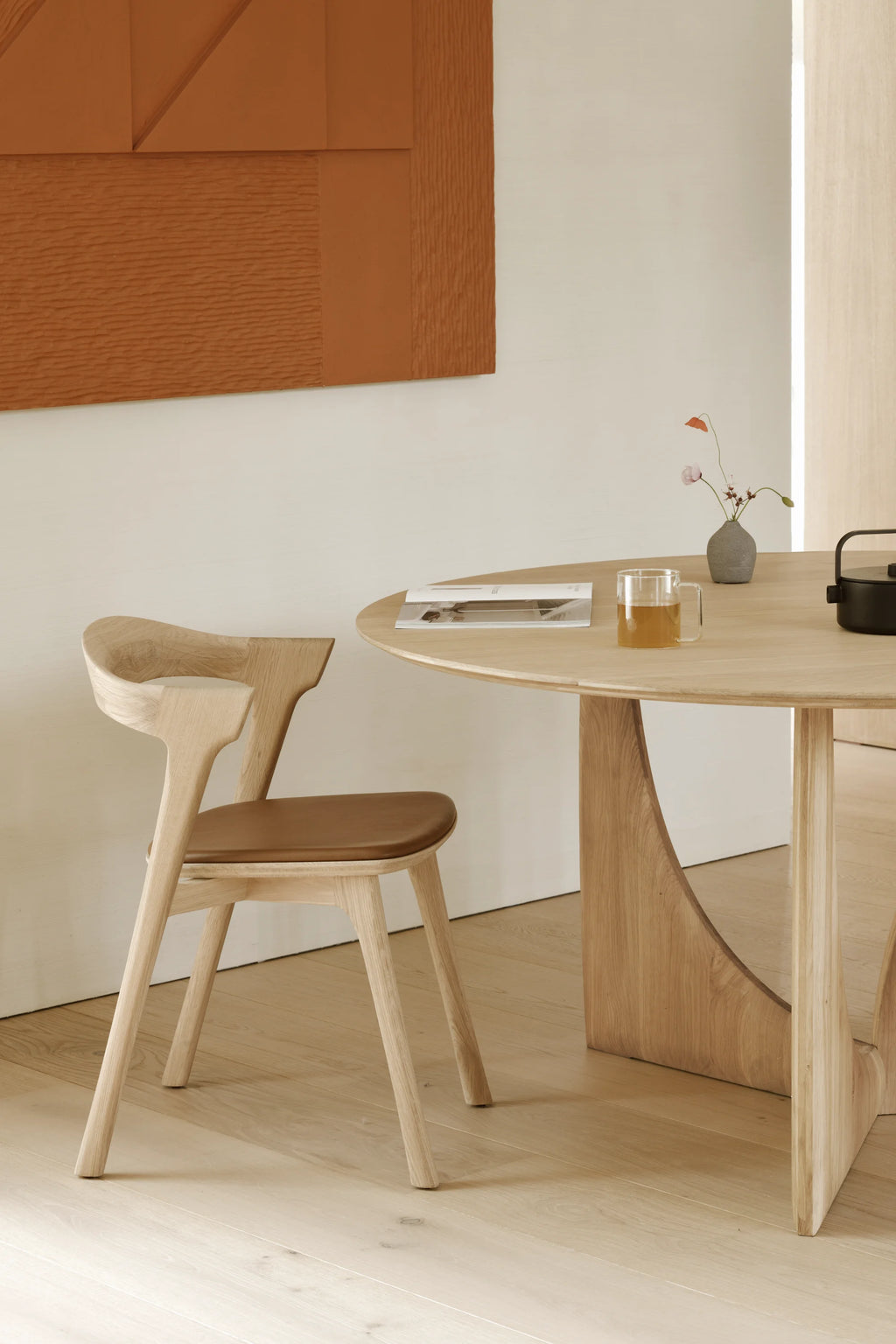 Ethnicraft Geometric Round Dining Table, a contemporary solid oak dining table featuring a smooth light natural oak finish and a distinctive sculptural geometric base with elegant curved lines. This modern round dining table offers a minimalist aesthetic and is perfect for Scandinavian dining rooms or organic modern kitchen spaces.