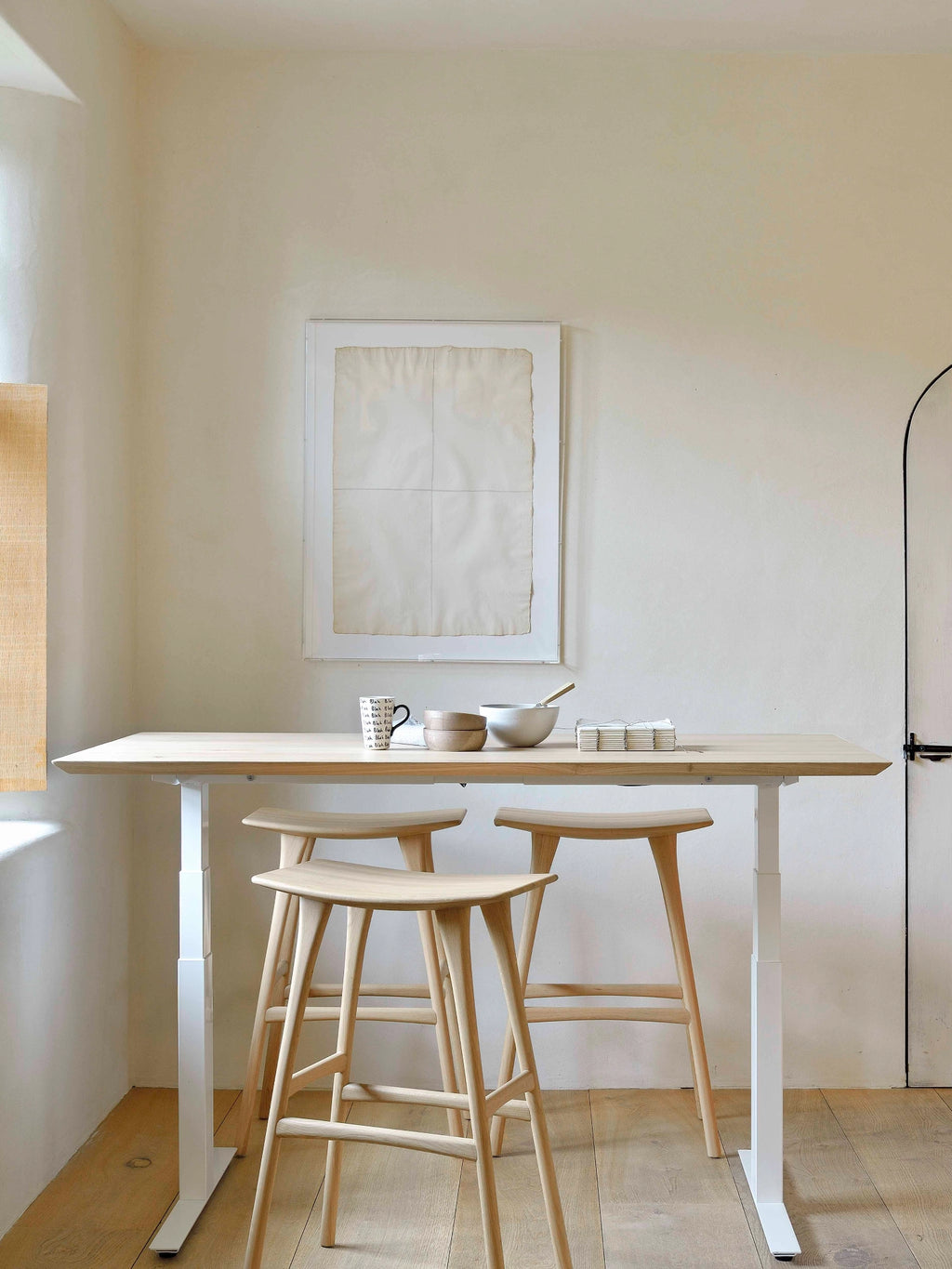 Ethnicraft Bok Adjustable Desk in natural oak with a light wood tabletop and a white adjustable frame, paired with two matching wooden bar stools in a bright, airy room.