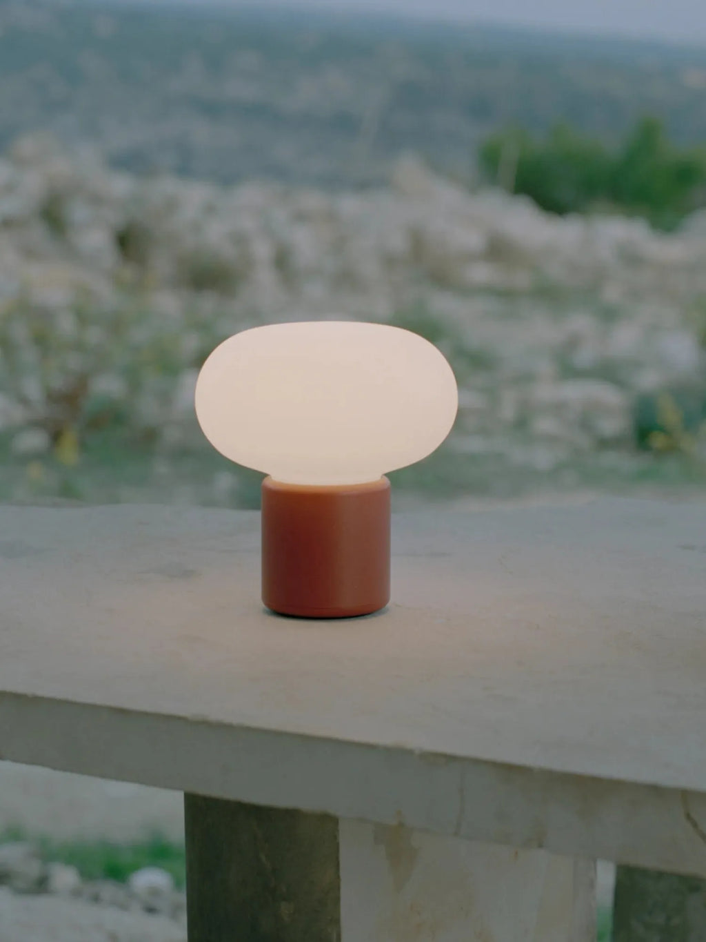 New Works Karl-Johan Portable Table Lamp with a matte burnt orange base and glowing white mushroom-shaped diffuser, displayed outdoors on a light concrete surface. Ideal for ambient lighting.