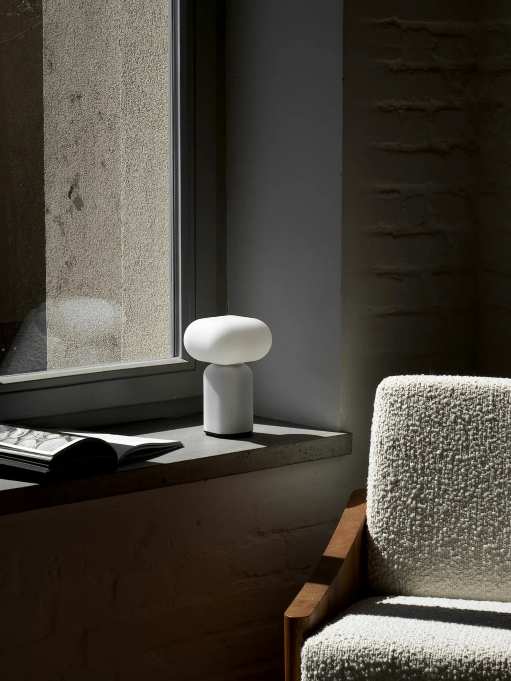TAMO Velis Portable Lamp, a minimalist white cylindrical lamp with an oval frosted light diffuser, sits on a sunlit concrete windowsill next to a book and a cozy cream textured armchair. A premium modern lighting solution.