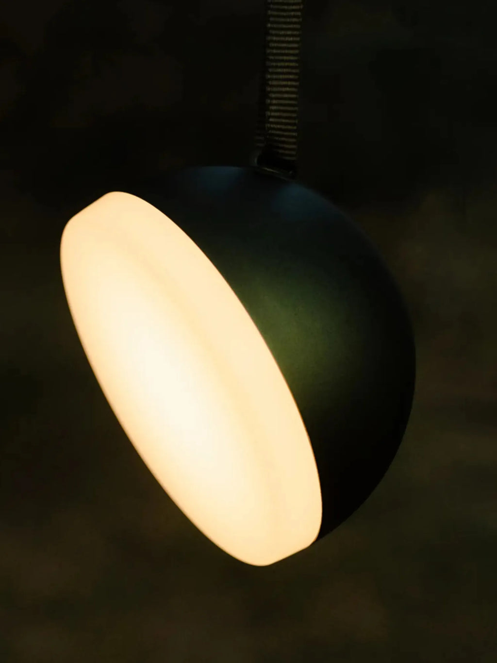 New Works Sphere Adventure Light, a black matte spherical portable lamp with a glowing warm white elliptical light cutout, hanging in a dark setting. This premium design is ideal for versatile ambient lighting.