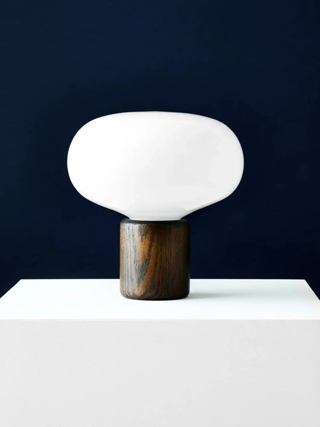 New Works Karl-Johan Table Lamp, featuring a smooth, matte white opal glass shade perched on a rich, dark brown wood base showcasing natural grain, displayed on a clean white surface against a deep blue background. A premium contemporary lighting addition.