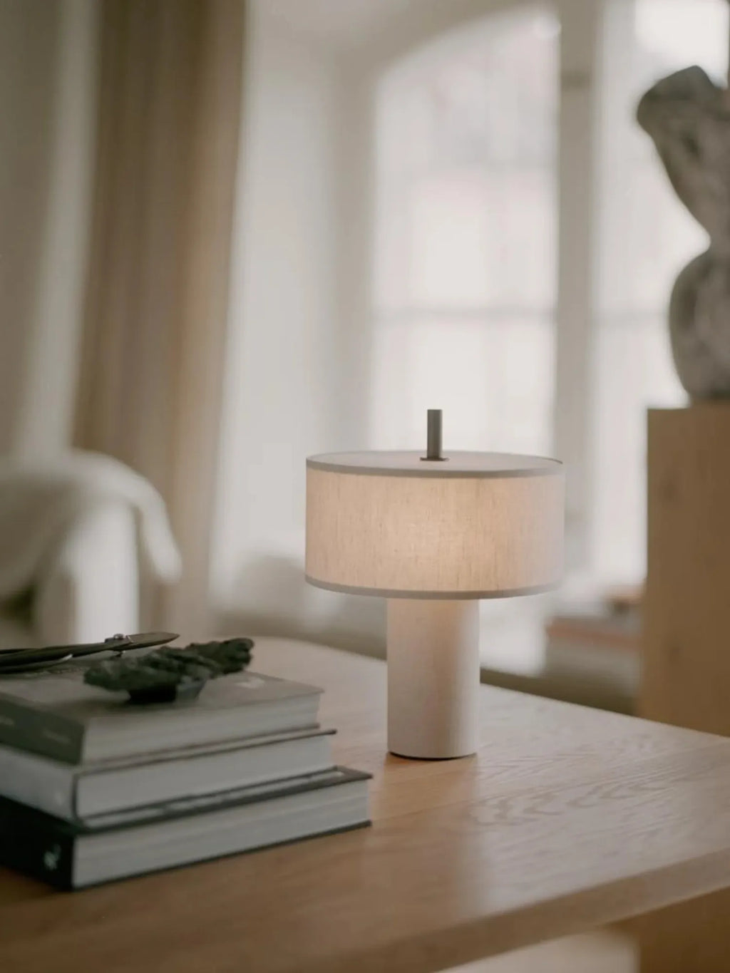 New Works Margin Portable Table Lamp, featuring a round, natural linen shade and a white cylindrical base with a dark brown top accent, on a light wooden table with books. A premium, modern lighting solution. Perfect for enhancing any contemporary space.