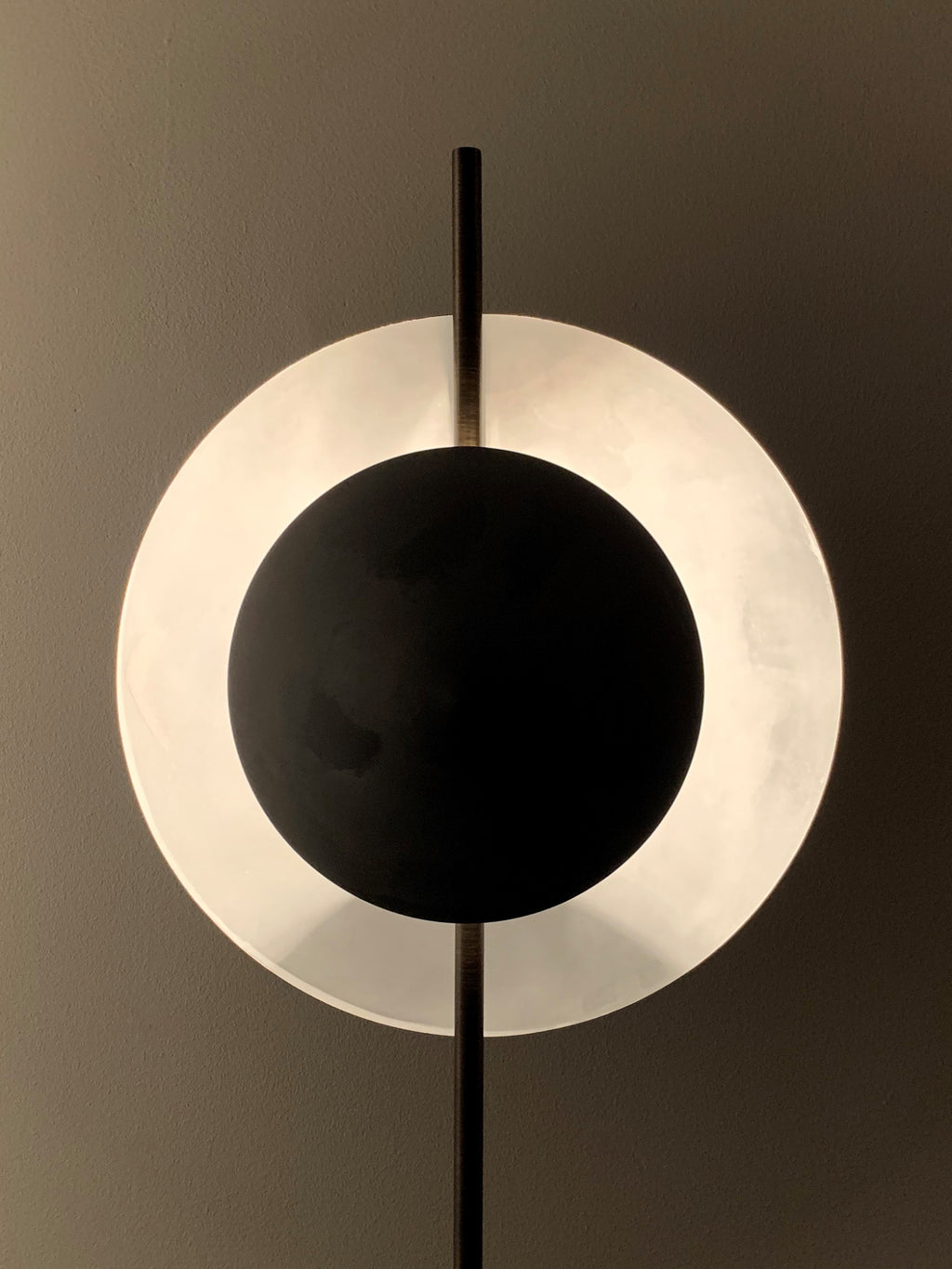 101 CPH Dawn Floor Lamp, featuring a unique circular design with a glowing translucent white outer ring surrounding a darker black matte inner disc, mounted on a dark vertical metal stand against a gray wall. This luxury accent lighting is ideal for modern interiors.