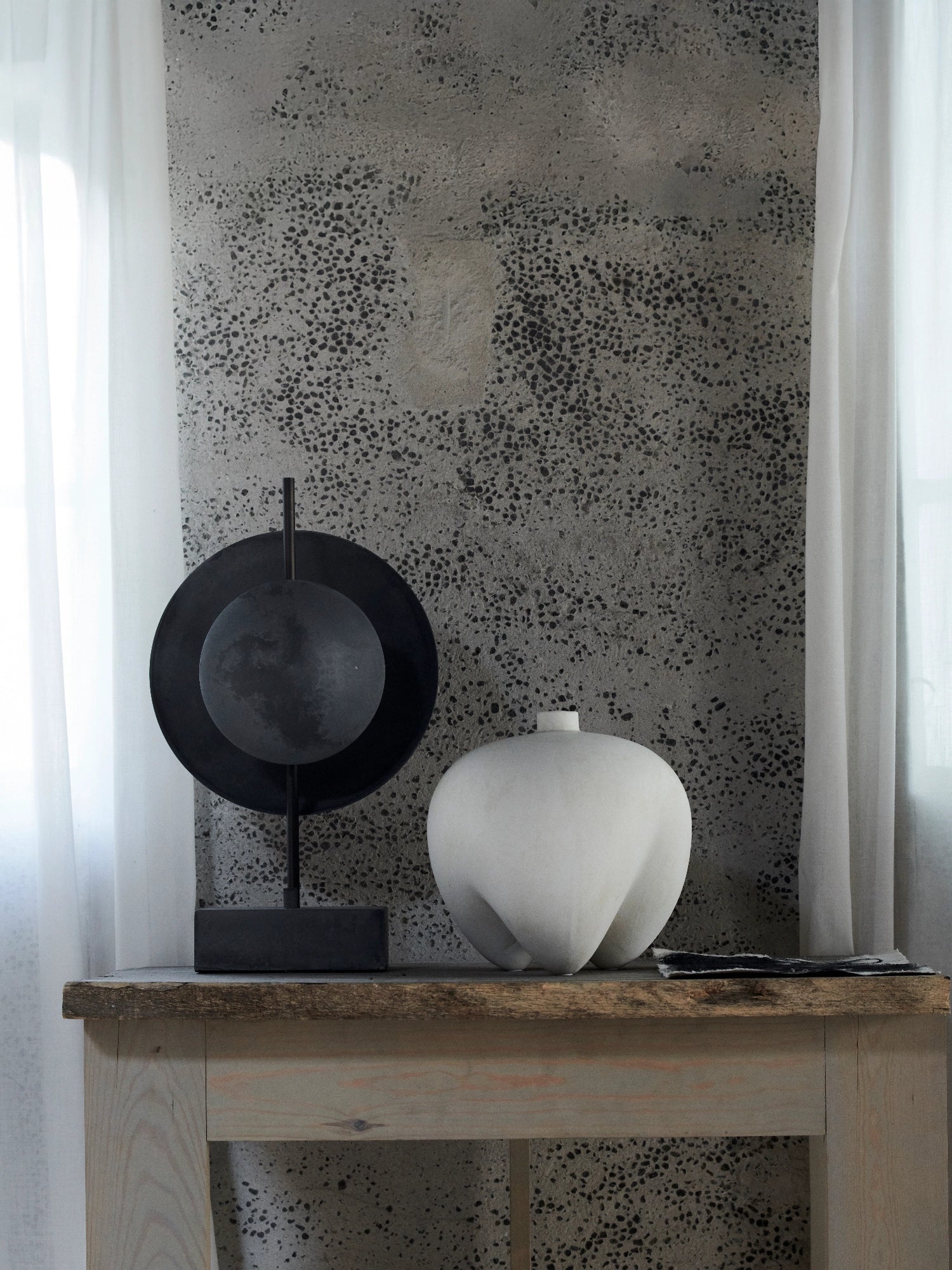 101 CPH Dusk Table Lamp features a matte black metal stand with a textured dark gray spherical "moon" element centered on a larger black disk, displayed on a rustic wooden table with a white ceramic vase. Ideal for creating a warm, ambient space.