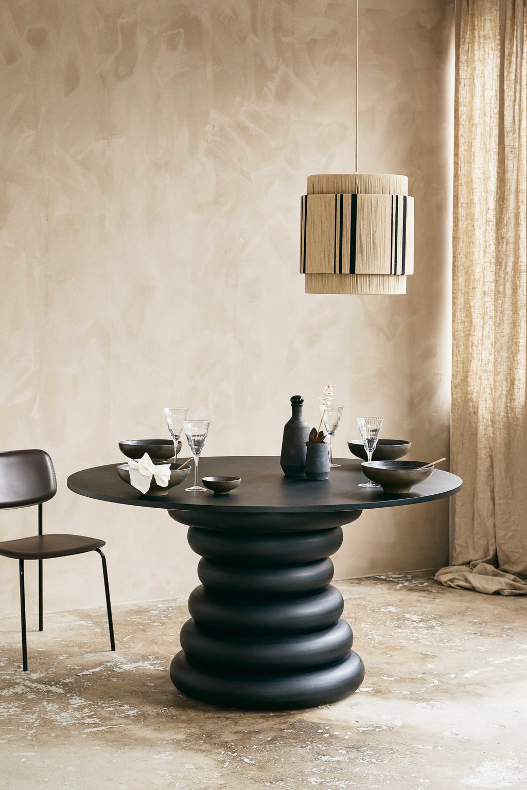 Nordal Yalu Dining Table, a striking matte black round dining table featuring a smooth, minimalist tabletop atop a bold, sculptural stacked ring base. This contemporary black dining table embodies an organic modern design with its unique silhouette and solid, grounding presence. Perfect as a statement dining table for modern minimalist or brutalist-inspired dining rooms and kitchens.