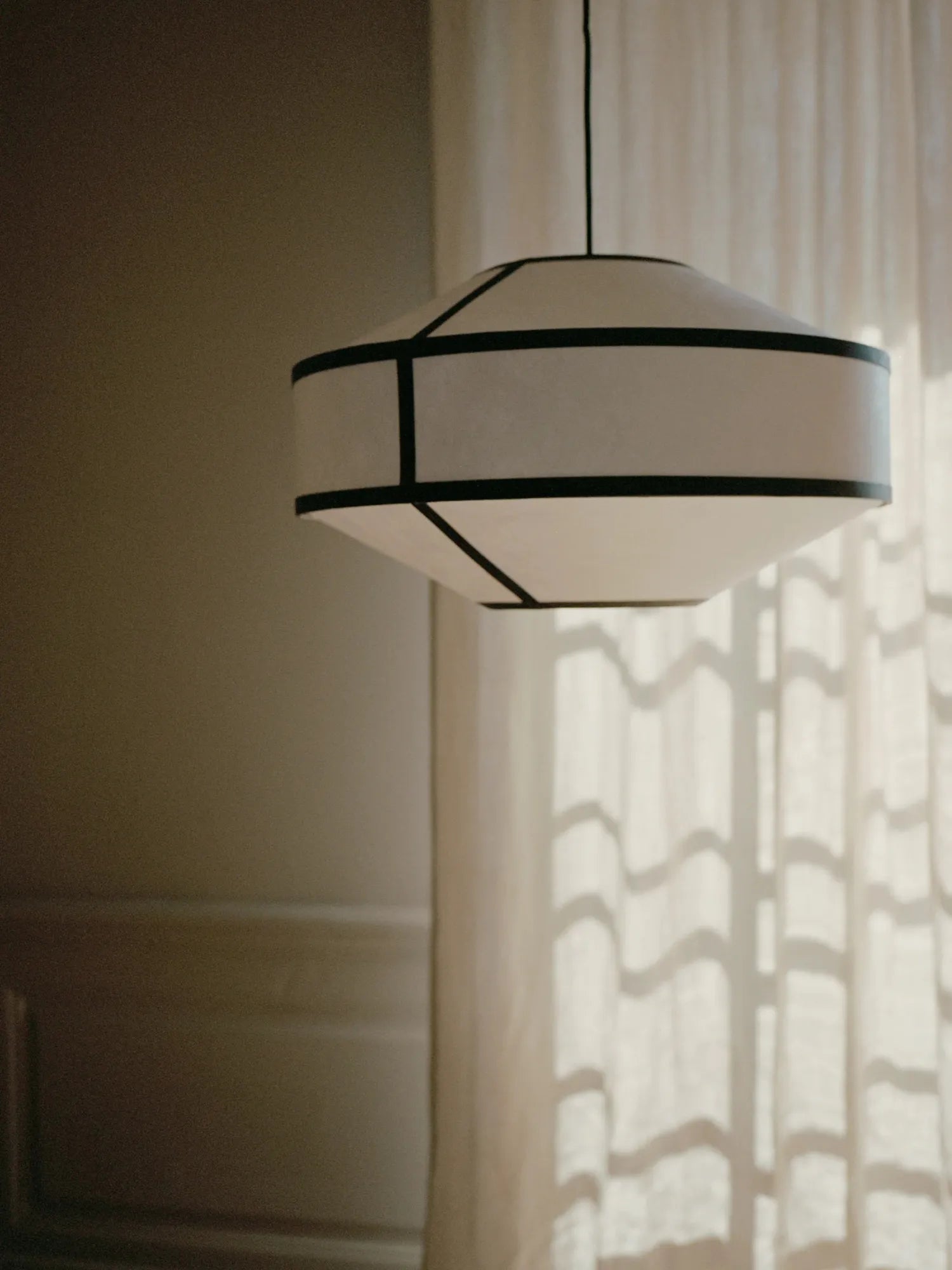 New Works Kite Pendant Lamp featuring a white paper shade with graphic black ribbon detailing, suspended against a softly lit window with sheer curtains. Offers premium, diffused light.