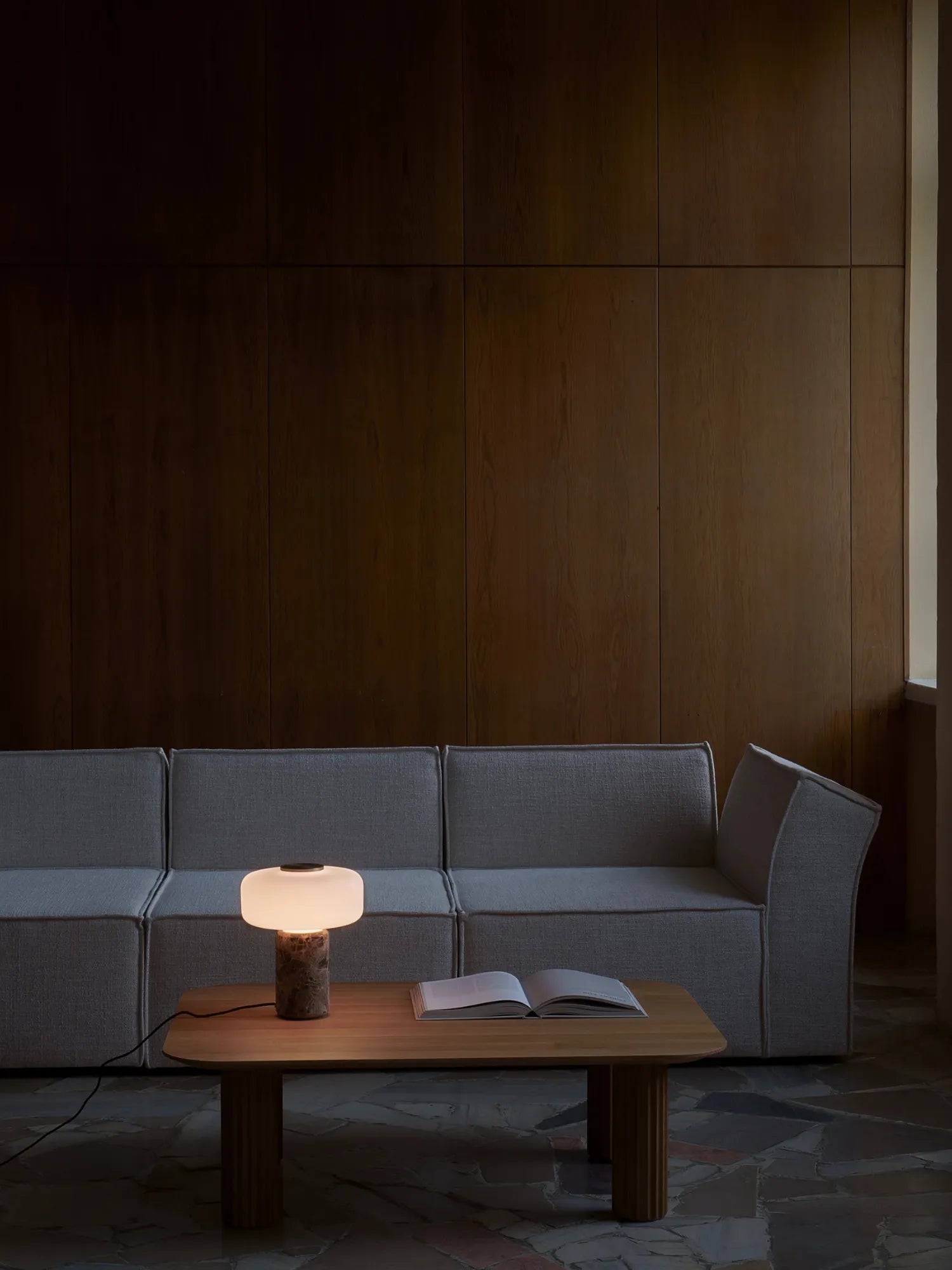 TAMO Fonte Table Lamp glowing on a wooden coffee table in a modern setting. This luxury desk lamp features a genuine marble base and an oblong frosted white glass shade, casting a warm ambient light. Perfect for sophisticated living spaces.