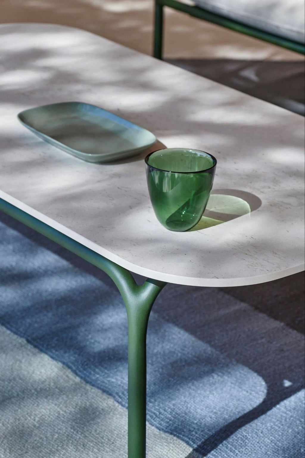 Gandia Blasco Capa Coffee Table, an organic modern outdoor coffee table featuring a light grey or off-white composite tabletop with softly rounded oval edges and a subtle textured finish. It stands on distinctive matte olive green metal legs with a branching, sculptural design, providing a contemporary aesthetic. Perfect for a stylish patio, garden, or outdoor living room setup, enhancing modern minimalist outdoor spaces.
