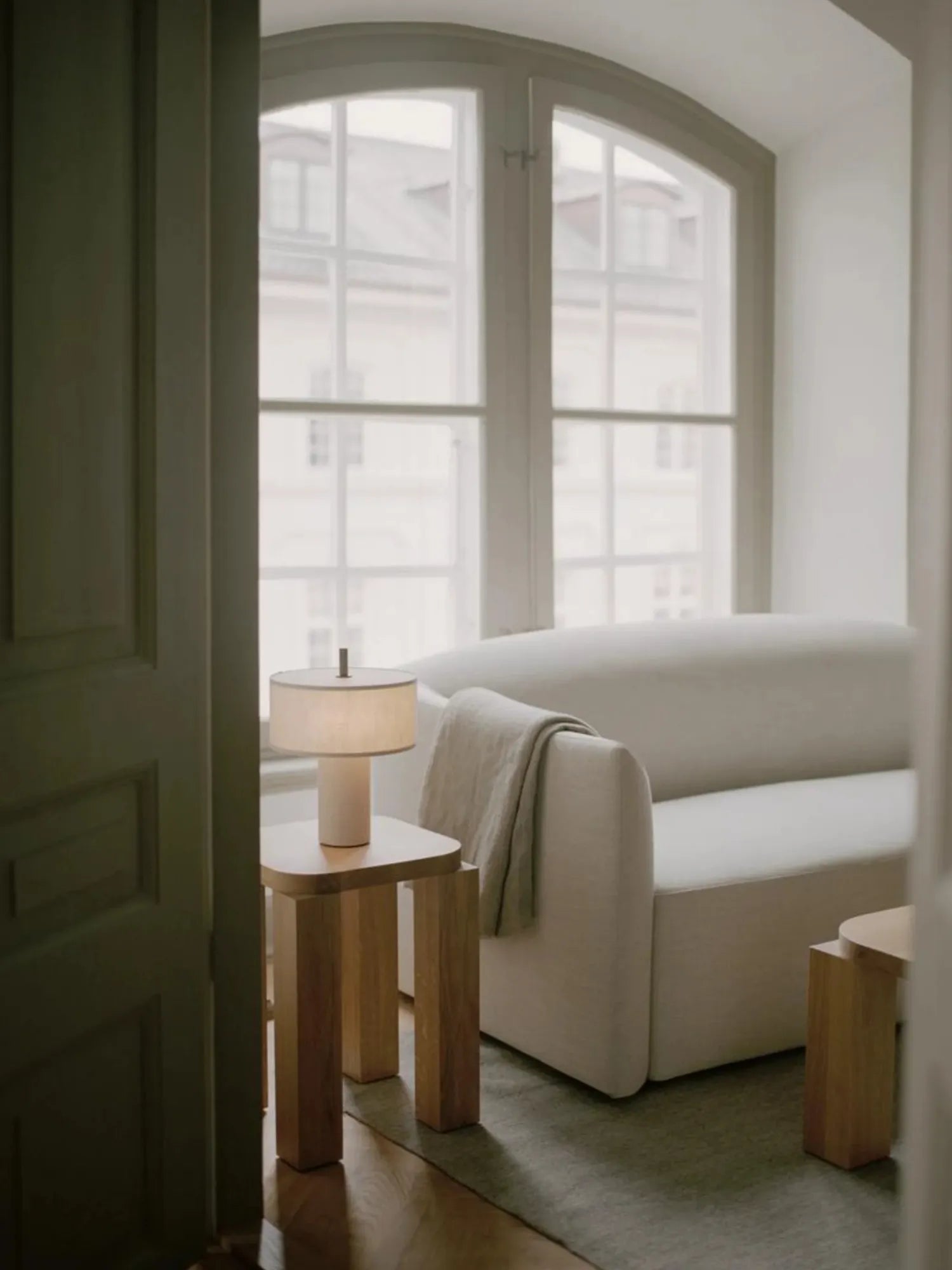 New Works Margin Portable Table Lamp, featuring a natural light-colored linen shade and a minimalist cream base, styled on a light-wood square side table in a cozy, cream living room. Ideal for creating inviting ambient lighting.