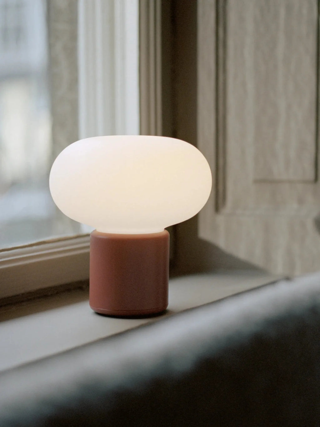 New Works Karl-Johan Portable Table Lamp with a glowing, soft white oval opal glass shade and a matte brownish-pink base, sitting on a window sill in a lifestyle setting. This luxury authentic lamp is perfect for adding ambiance.