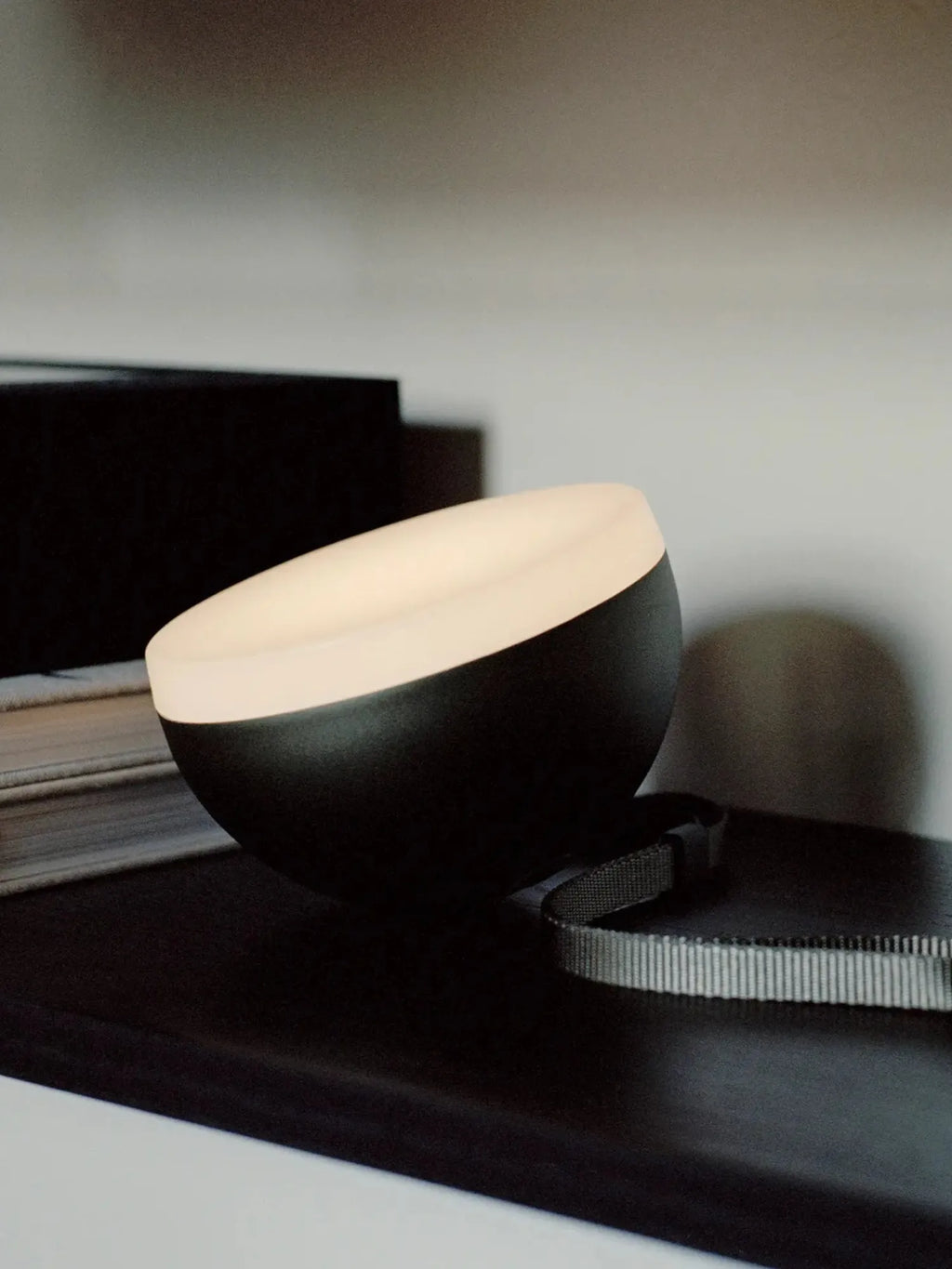 New Works Sphere Adventure Light, a modernist portable lamp with a matte black spherical base and luminous frosted white dome, featured on a shelf next to books. Ideal for contemporary interior lighting.