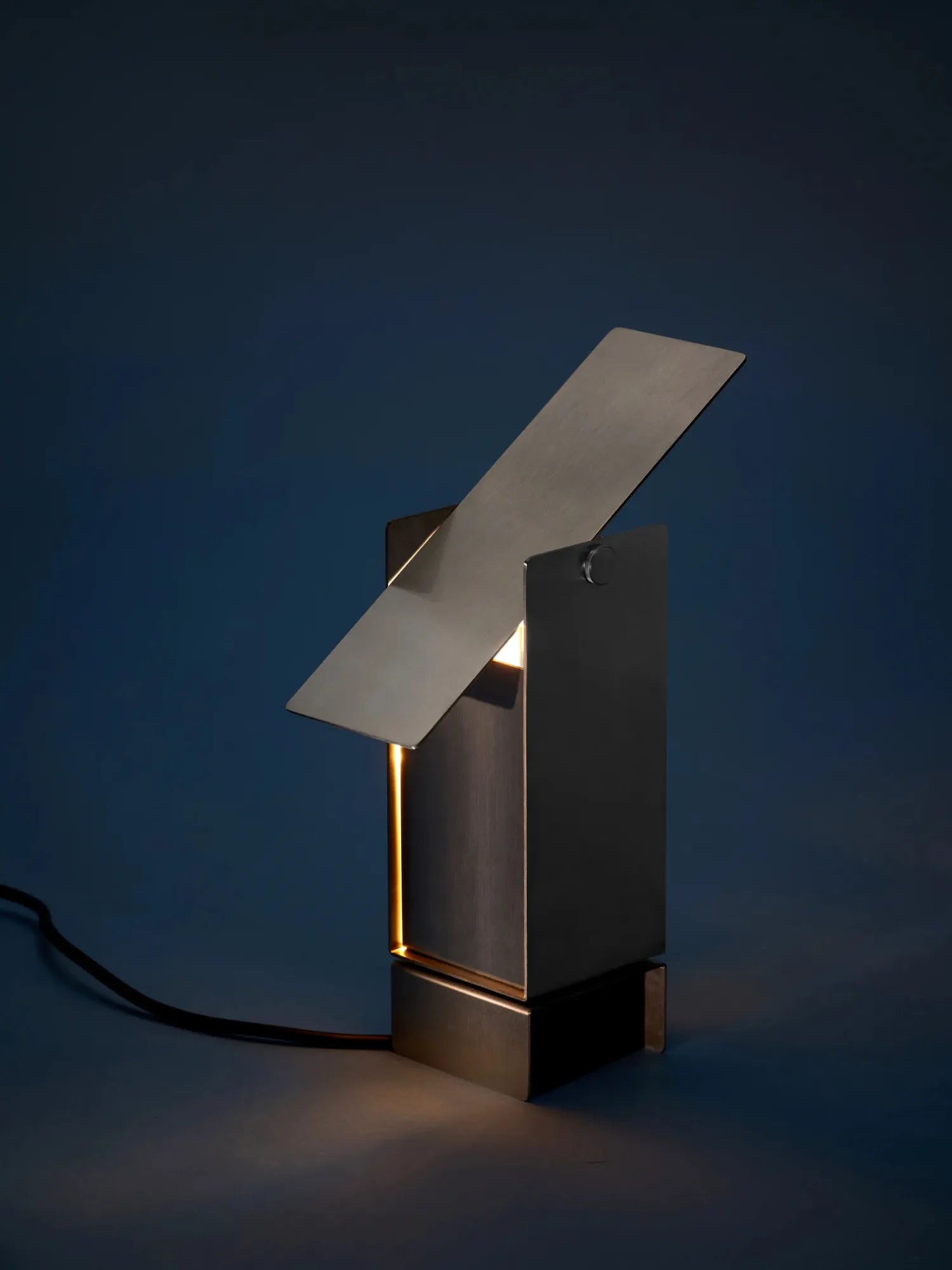 MOEBE Pivoting Table Lamp, a modern minimalist desk lamp crafted from dark anthracite brushed steel with a striking geometric silhouette and an adjustable pivoting top panel. This industrial chic table lamp features a warm, directional light source from a hidden slot, allowing for precise illumination. Perfect as a contemporary bedside lamp, home office desk lamp, or accent lighting for Scandinavian and industrial interiors.