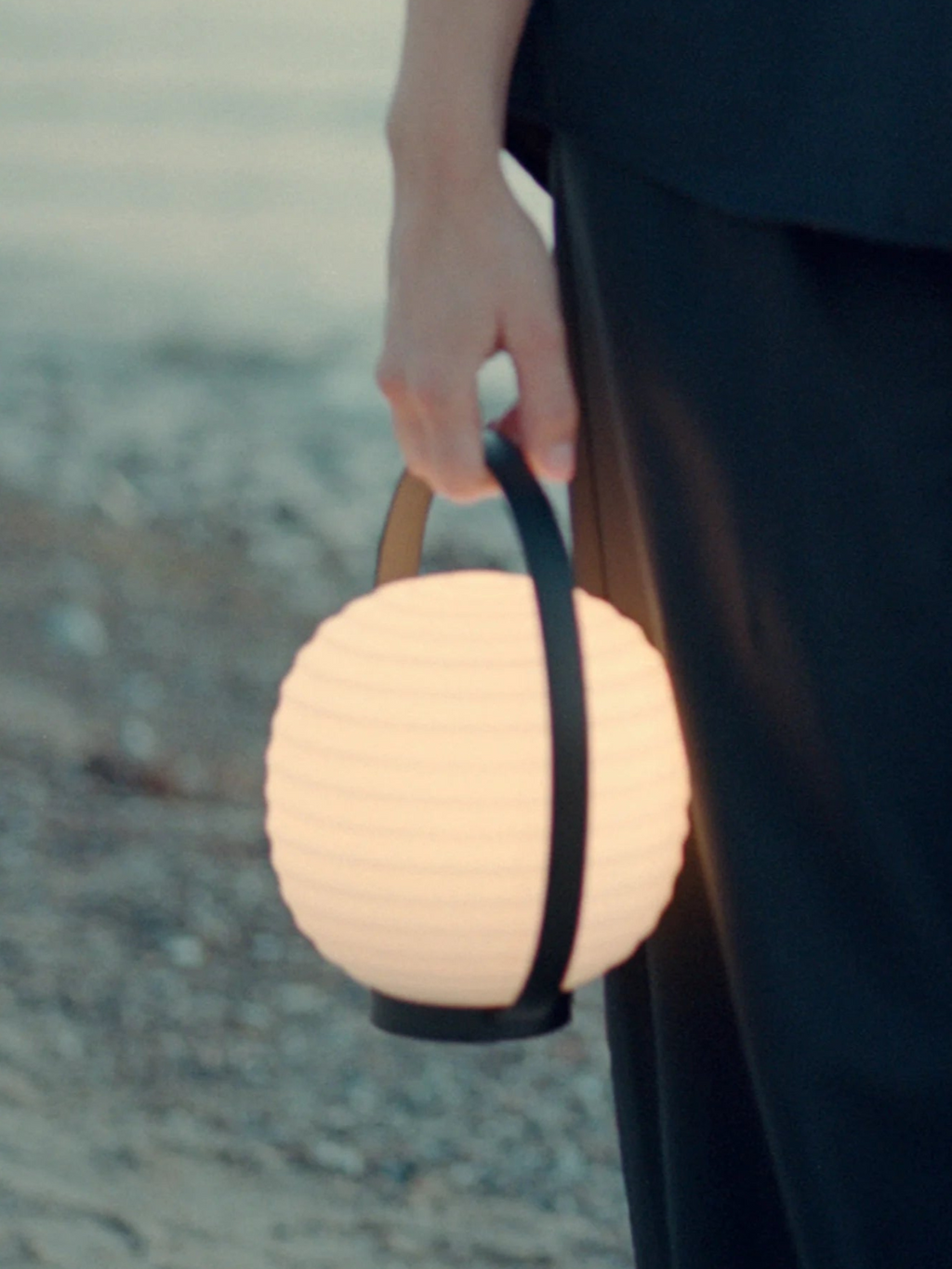 New Works Lantern Portable Lamp held in a hand, featuring a glowing white spherical shade with horizontal ridges and a black leather-like handle and base. This modern, luxury lamp is perfect for ambient indoor or outdoor lighting.
