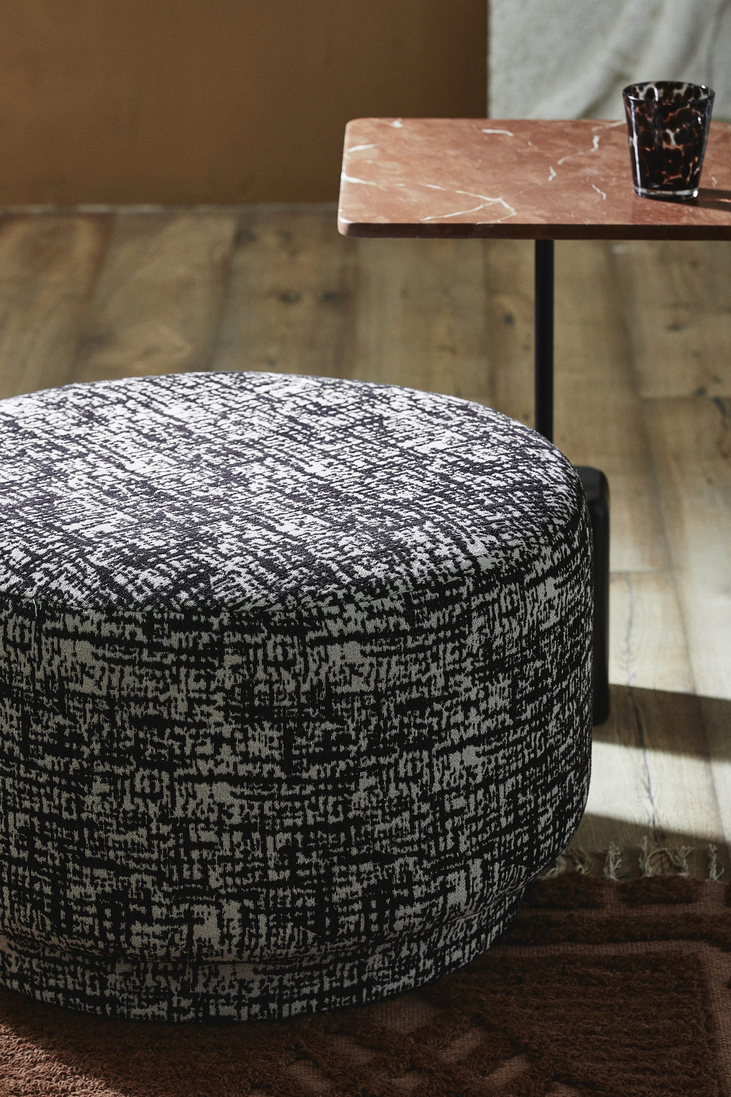 Nordal Bomu Pouf, a cylindrical round pouf upholstered in a textured black and white fabric with an abstract, graphic pattern. Its contemporary design features an intricate woven texture that creates a dynamic visual interest, offering both comfort and modern style. This versatile accent ottoman is perfect for modern living rooms, bedrooms, or as stylish extra seating in any contemporary space.