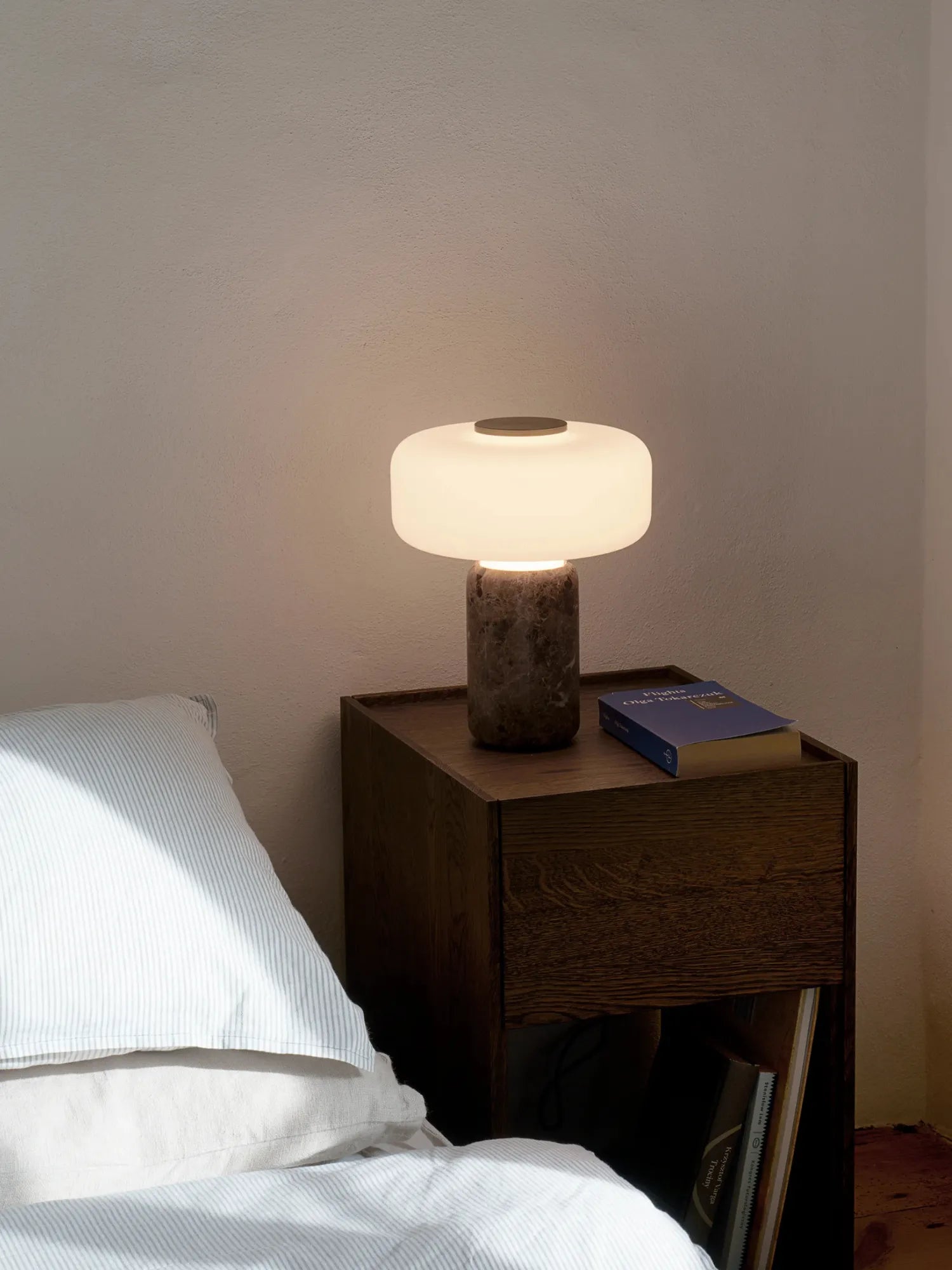 TAMO Fonte Table Lamp, featuring a luminous white glass shade atop a premium dark brown marble base, glowing softly on a nightstand. Perfect for ambient bedroom lighting.
