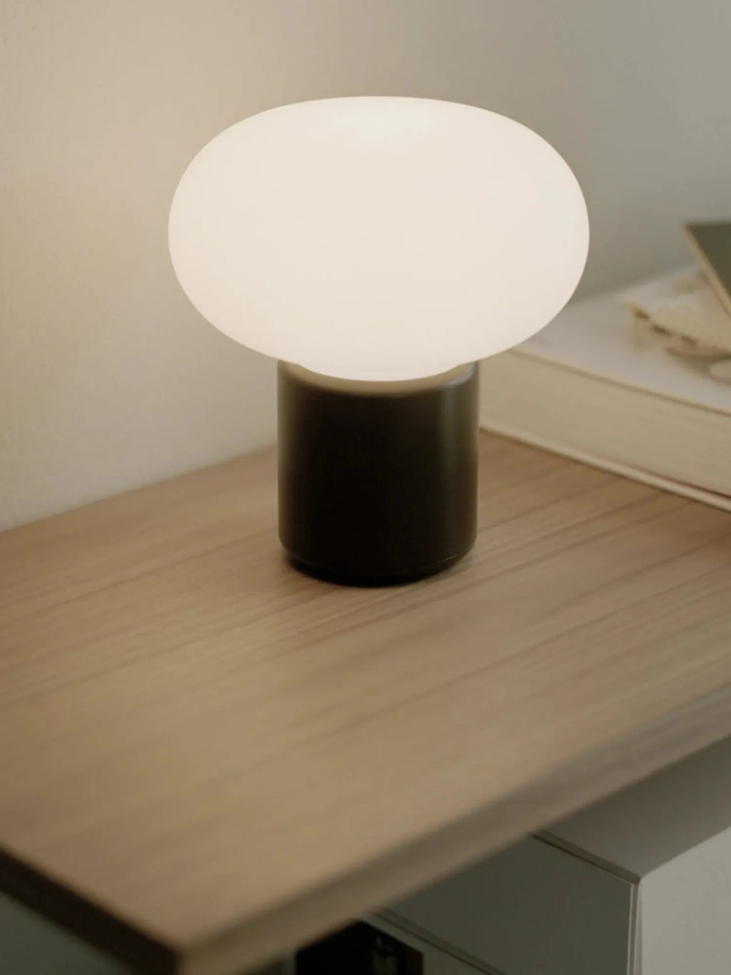 New Works Karl-Johan Portable Table Lamp with a luminous white opal glass sphere atop a dark, matte brown base, pictured on a light wood bedside table with a book. A luxury lighting solution for any setting.