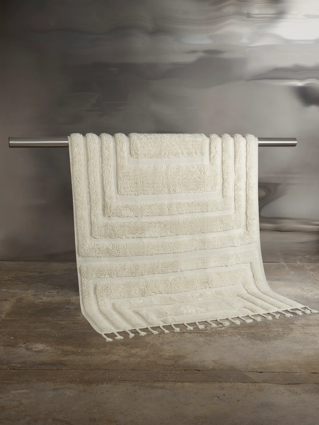 Tinted Objects Kask Wool Rug, a luxurious cream-colored wool rug with a textured geometric pattern and fringe detailing. This soft and stylish floor covering is perfect for adding warmth and elegance to any modern living space or bedroom.