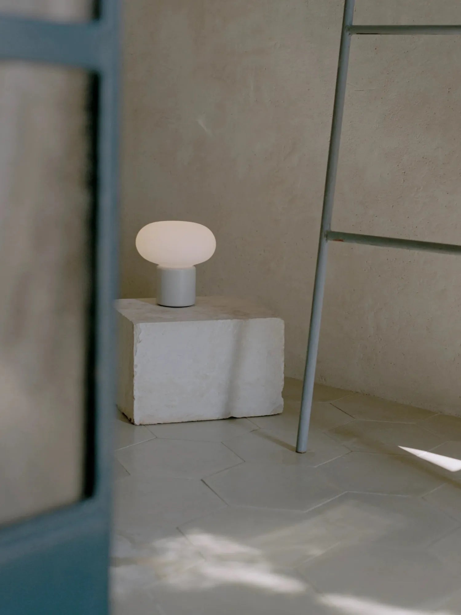 New Works Karl-Johan Portable Table Lamp with a milky white mushroom-shaped glass shade and a light gray cylindrical base, displayed on a textured white plinth in a minimalist room. This modern, premium LED lamp provides ambient portable lighting.