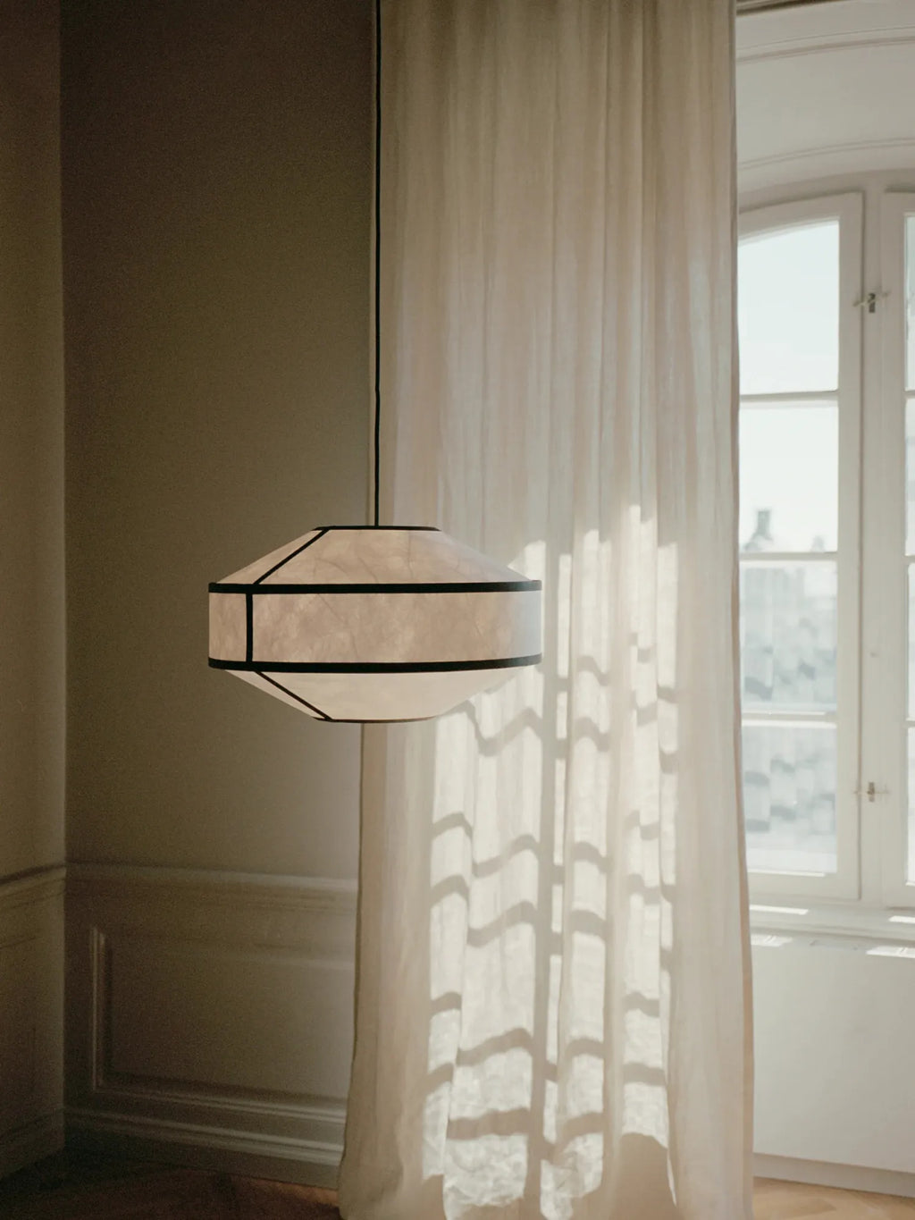 New Works Kite Pendant Lamp featuring a luxury cream rice paper shade with black accent trim, hung in a sunny room with white curtains. Ideal for modern Scandinavian interiors.