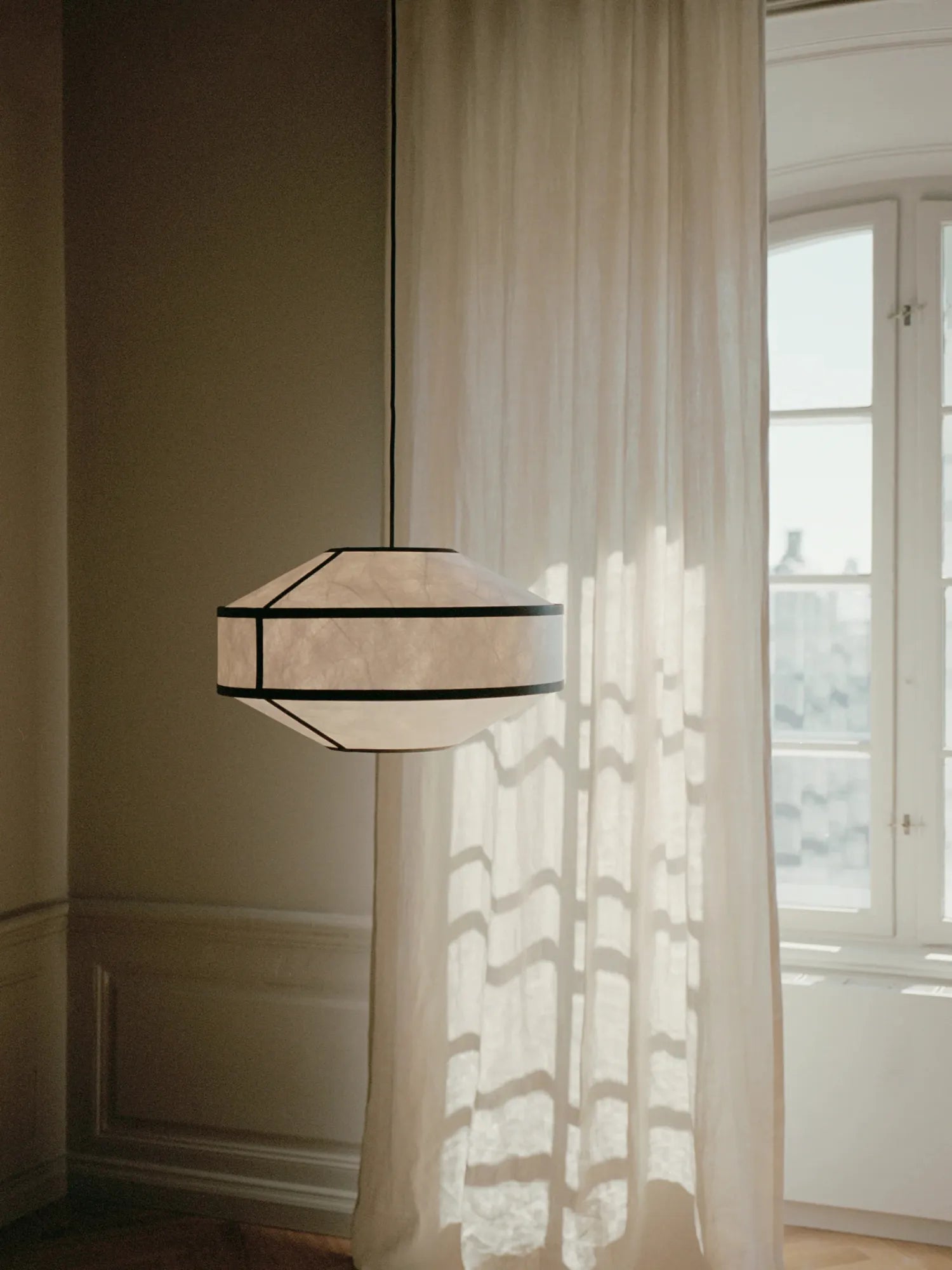 New Works Kite Pendant Lamp featuring a luxury cream rice paper shade with black accent trim, hung in a sunny room with white curtains. Ideal for modern Scandinavian interiors.