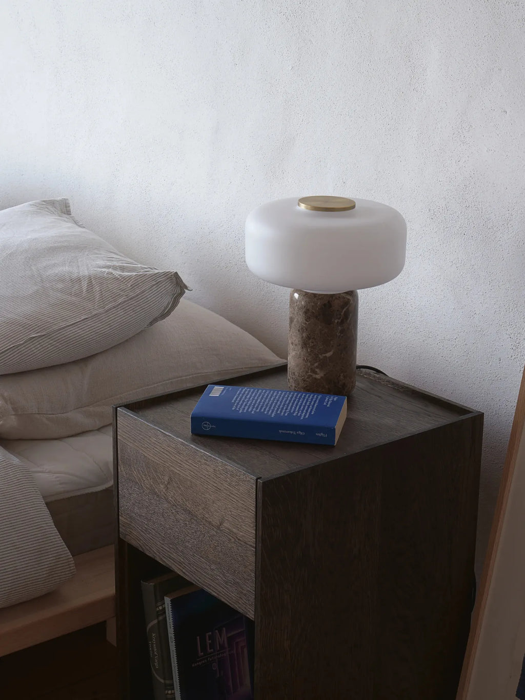 TAMO Fonte Table Lamp: A luxurious modern lamp with an opaque white glass diffuser, a polished brass top accent, and a round grey-brown marble base, displayed on a dark wood nightstand. Perfect for elegant bedroom lighting.