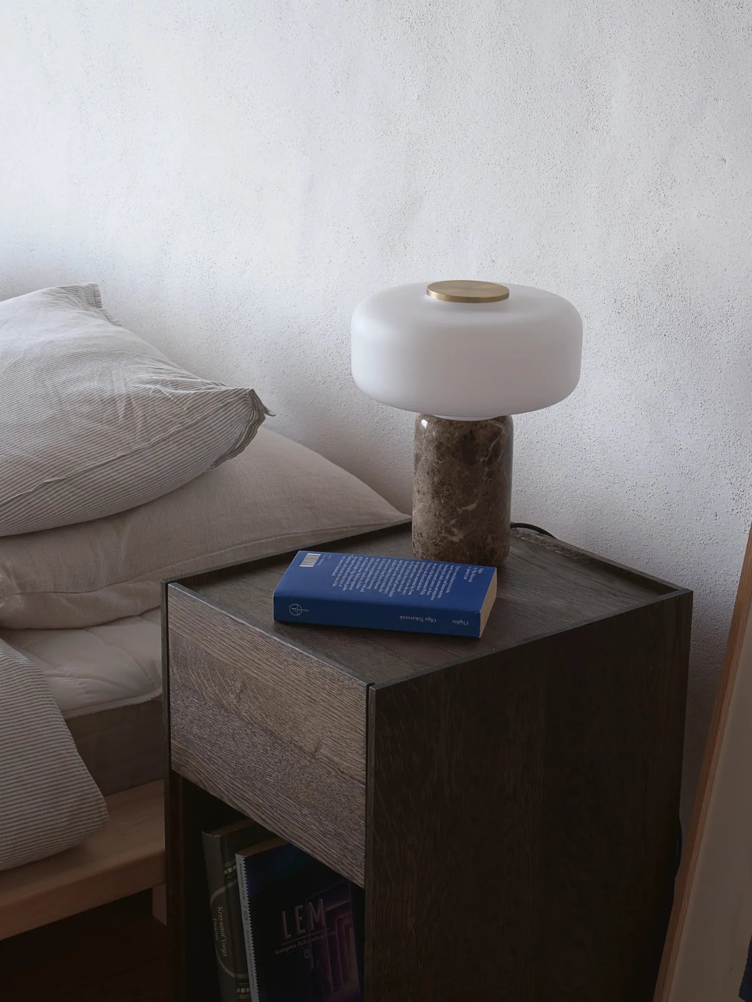 TAMO Fonte Table Lamp: A luxurious modern lamp with an opaque white glass diffuser, a polished brass top accent, and a round grey-brown marble base, displayed on a dark wood nightstand. Perfect for elegant bedroom lighting.