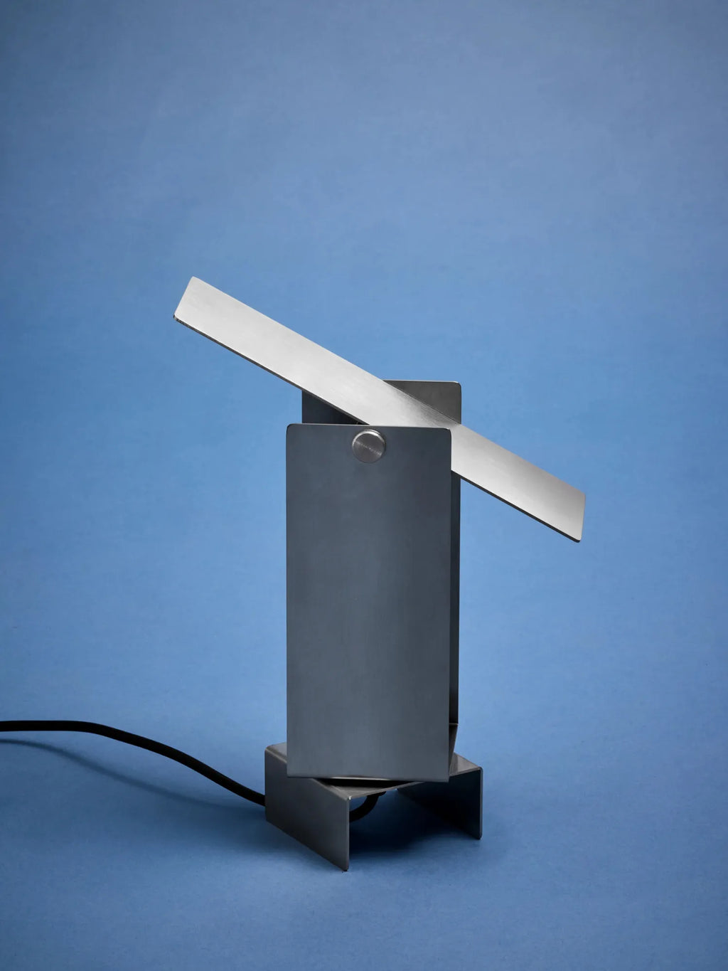 MOEBE Pivoting Table Lamp, a modern minimalist table lamp crafted from matte grey brushed stainless steel with a distinctive geometric, brutalist-inspired design. It features a unique pivoting rectangular panel that allows for adjustable light direction, secured by a visible circular screw. Ideal as an industrial chic desk lamp for a home office, a contemporary bedroom accent, or a minimalist living room light.