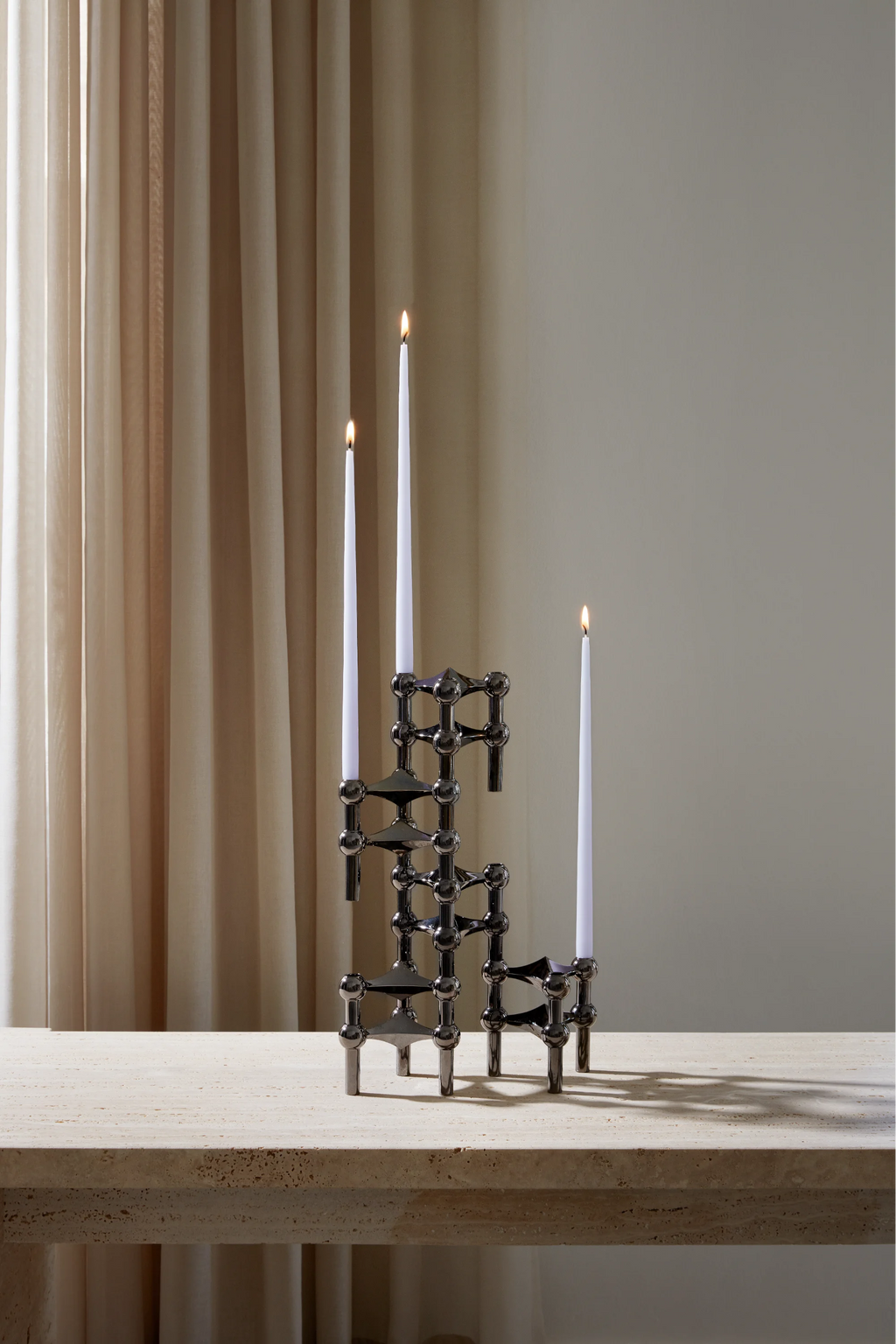 Stoff Nagel Candle Holder in a sleek black chrome finish, configured into an asymmetrical, multi-tiered structure holding three lit white taper candles, placed on a textured light beige stone table.