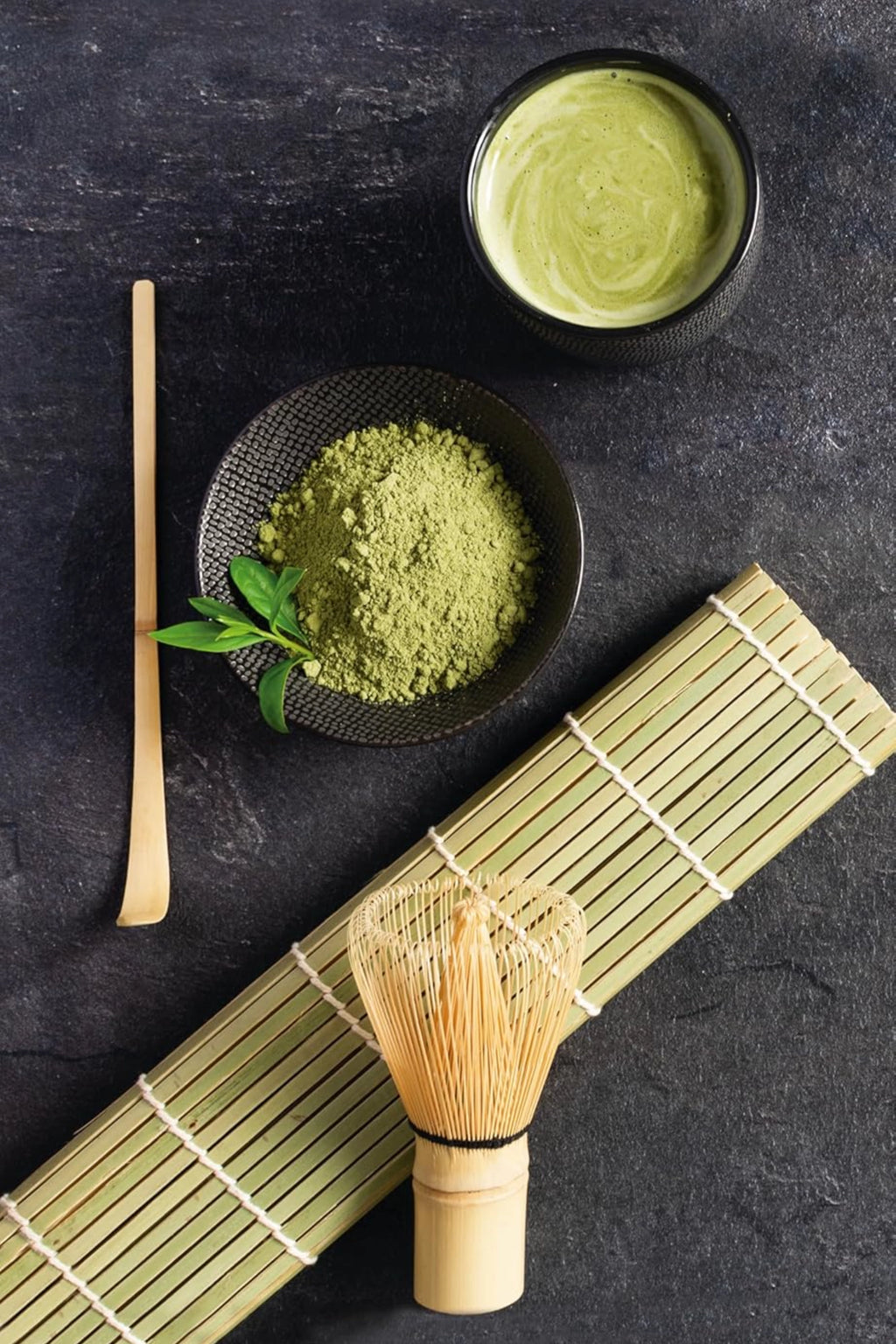 Ibili Matcha Set, a complete Japanese matcha tea ceremony kit featuring vibrant green ceremonial grade matcha tea powder and a foamy matcha latte in dark textured ceramic chawan bowls.