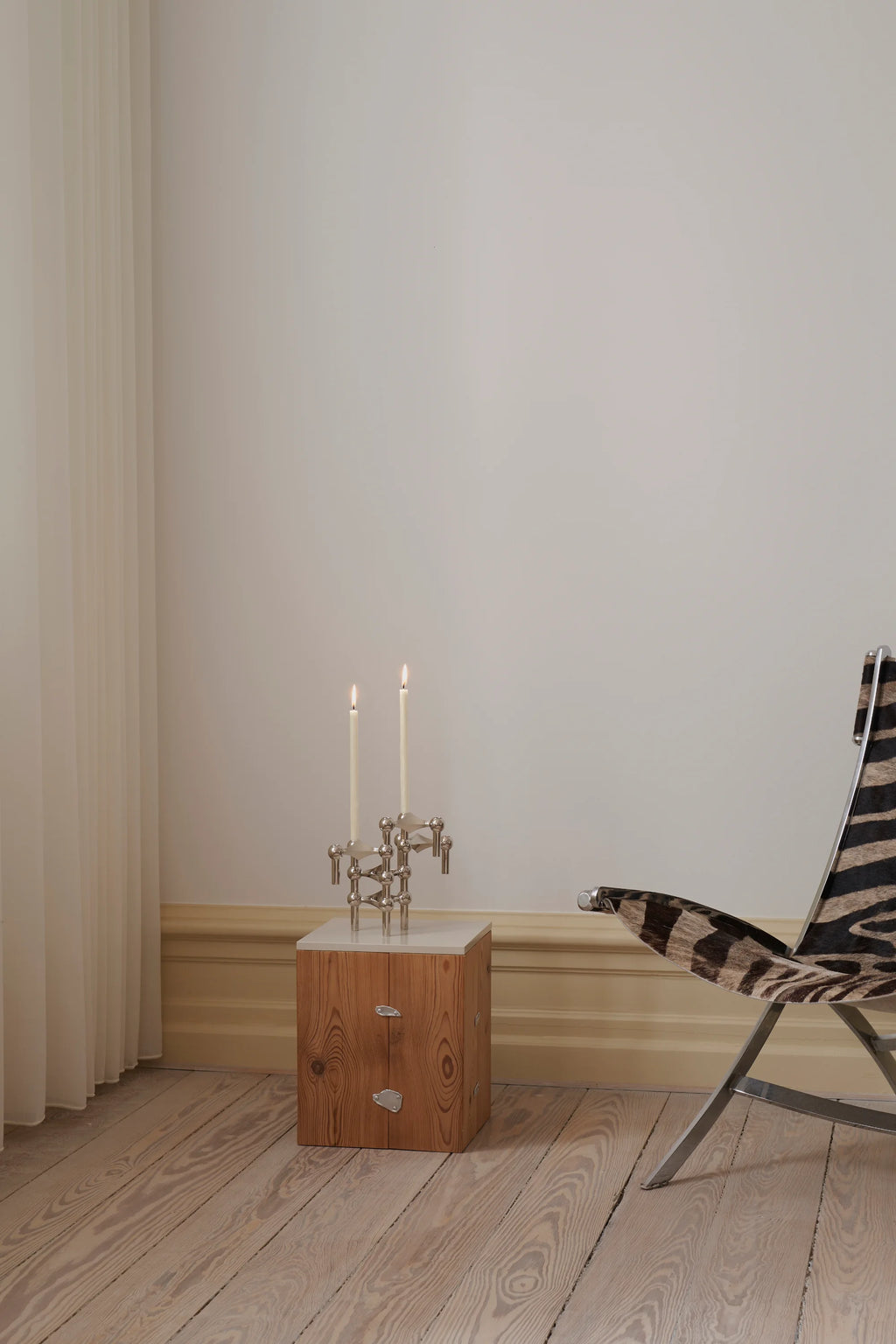 Stoff Nagel Candle Holder in a polished chrome finish, arranged to hold two lit cream taper candles, sitting atop a light wooden side table with a white marble top, next to a stylish zebra-print chair.