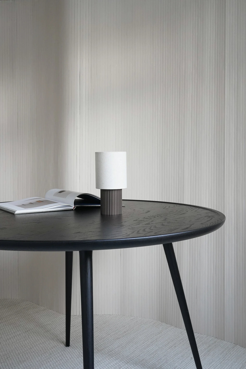 Mater Accent Round Dining Table, featuring a dark stained oak top with visible wood grain and sleek, tapered legs, styled with a modern white and dark gray table lamp and an open book, against a textured white wall.