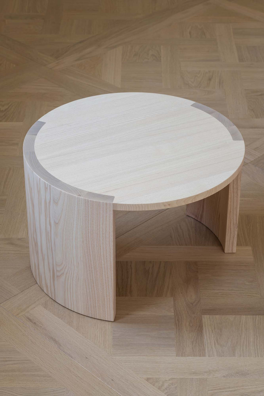 Made by Choice Airisto Coffee Table, a round modern minimalist coffee table crafted from light natural ash wood, featuring a smooth circular top and a unique sculptural base formed by two curved, semi-cylindrical panels that support the tabletop. The pale wood grain adds subtle texture and warmth, embodying organic modern and Scandinavian design principles. Perfect as a stylish centerpiece for contemporary living rooms or cozy minimalist spaces.
