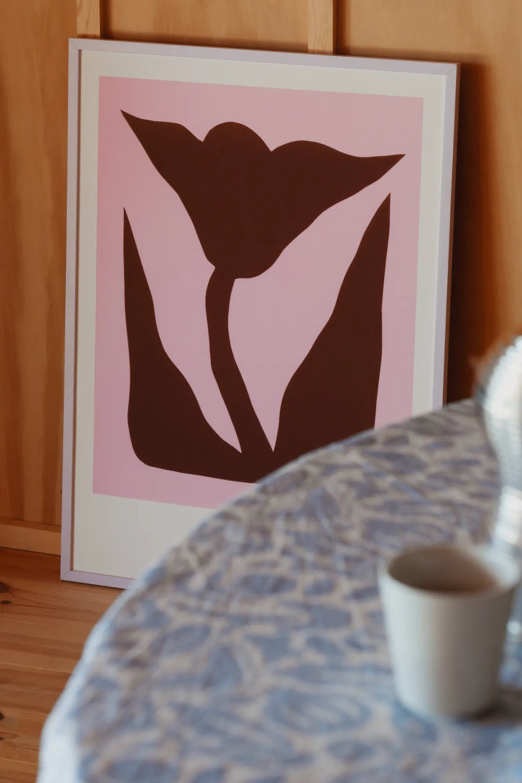 The Poster Club Mountain Lily, a modern abstract art print featuring a bold dark brown organic flower silhouette against a soft light pink background, framed with a crisp white mat and a subtle light lilac frame. This minimalist botanical poster is perfect for contemporary gallery walls, bedroom decor, or adding a touch of organic modern style to living rooms.