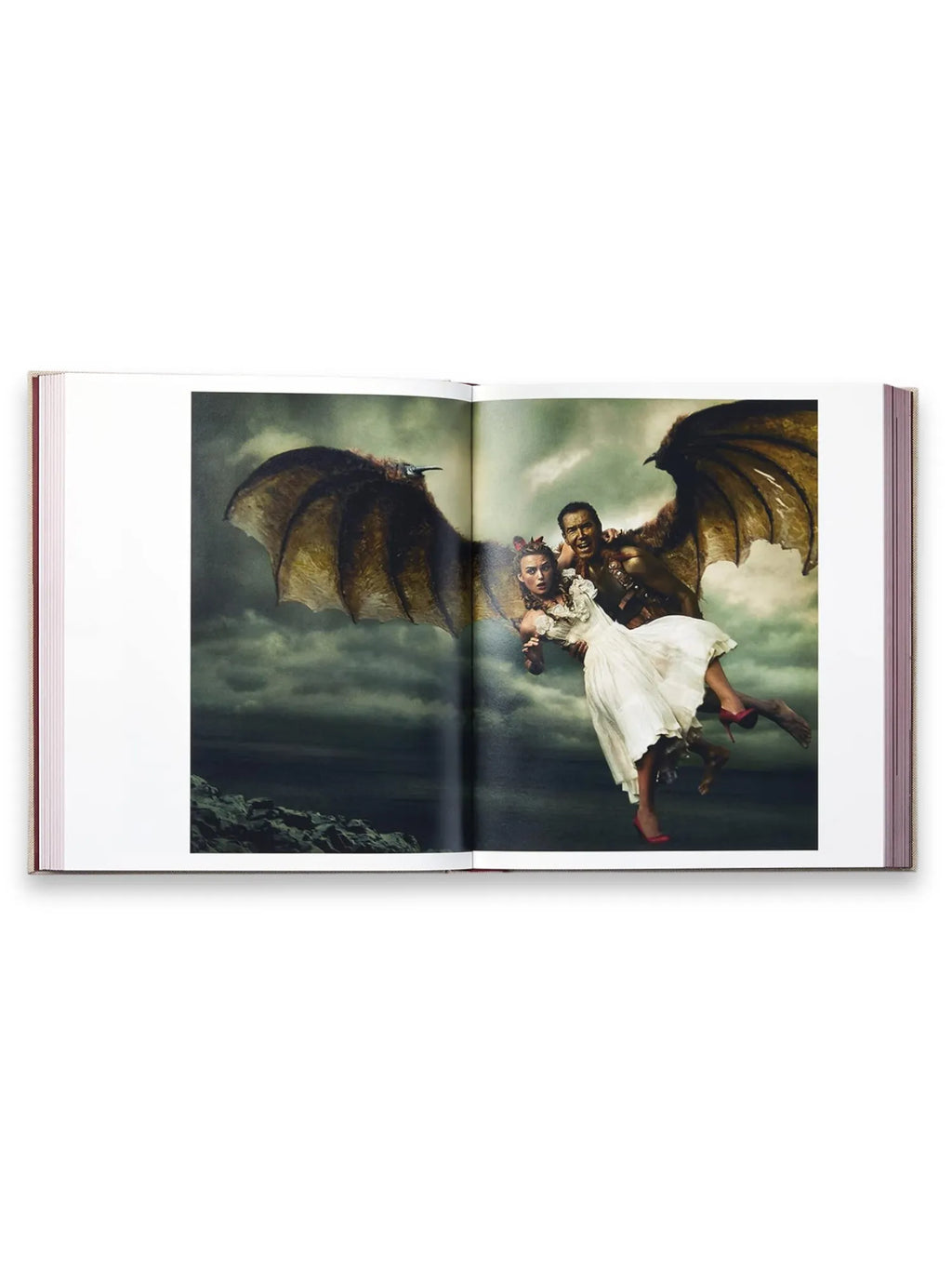 Book Annie Leibovitz’s ‘Wonderland’, an open photography book displaying a dramatic, vibrant full-page spread featuring a costumed man with large, dark wings carrying a woman in a flowing white dress and red heels against a stormy sky background. This collectible art book is perfect for photography enthusiasts and fans of Annie Leibovitz's iconic work.