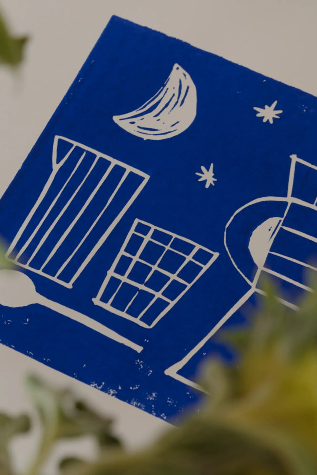 The Poster Club Aube, a striking abstract art print featuring bold white linework on a vibrant royal blue background, depicting a crescent moon, stars, and graphic architectural shapes with a handcrafted, textured block print.
