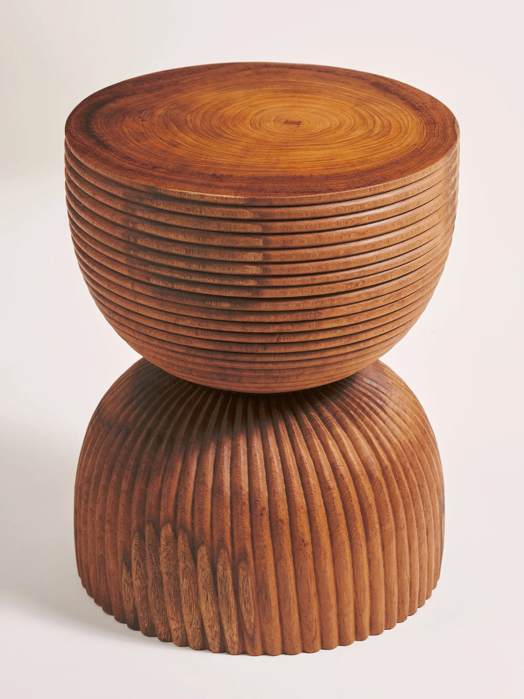 Heaps & Woods Anuk Stool, a sculptural solid wood stool crafted from warm brown wood with a rich grain, featuring a unique hourglass silhouette. The top half showcases concentric horizontal grooves, while the base features vertical undulating carved textures, creating a brutalist-inspired organic modern aesthetic. This versatile accent table is perfect for a contemporary living room, bedroom, or as a distinctive plant stand.