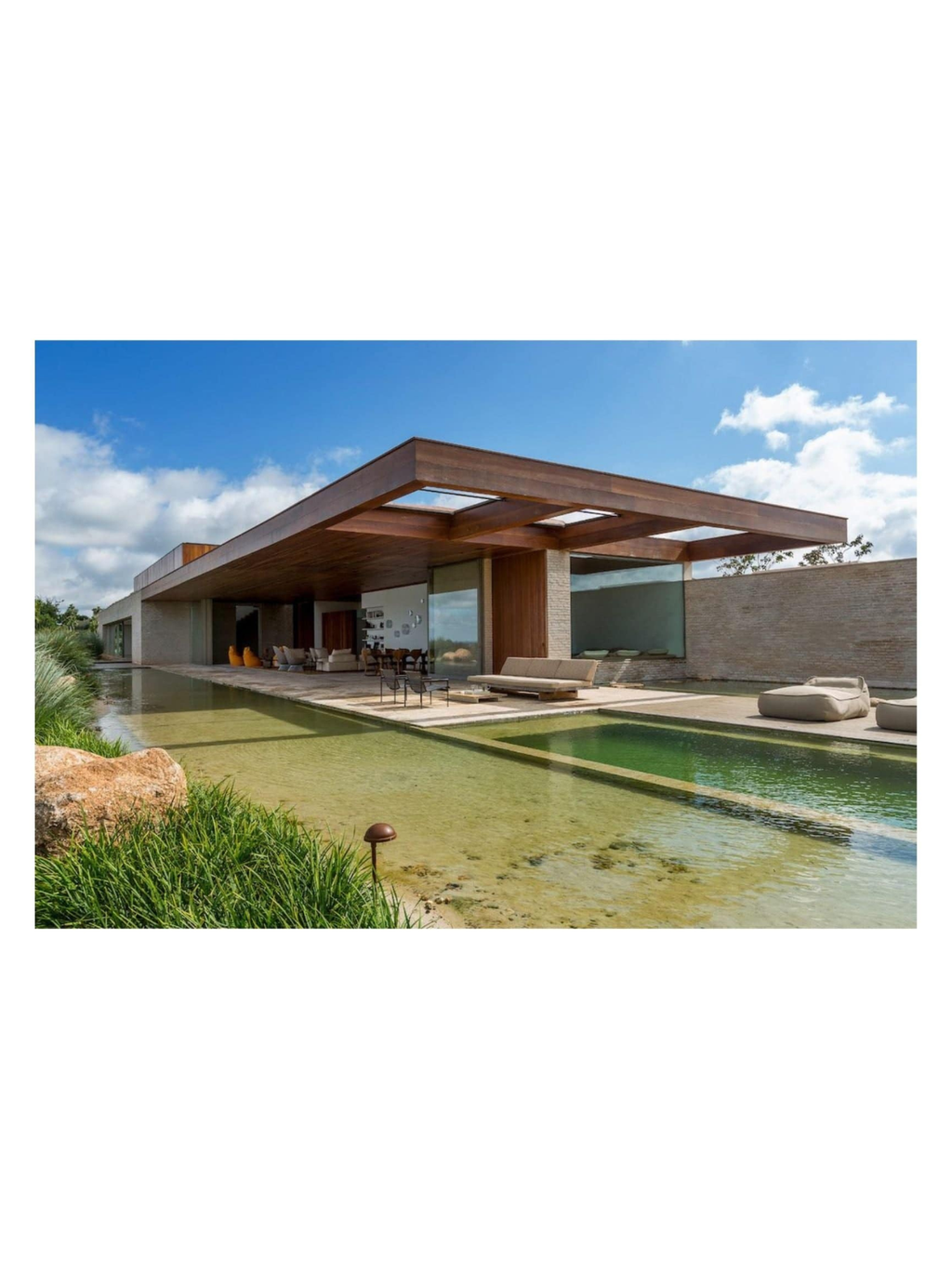 Book Arthur Casas: Architecture, featuring a luxurious modern home with a flat wooden roof, large glass walls, and an expansive outdoor living area with a naturalistic pool and lush green landscaping under a bright blue sky. This premium architecture book is a must-have for design enthusiasts and professionals seeking inspiration.