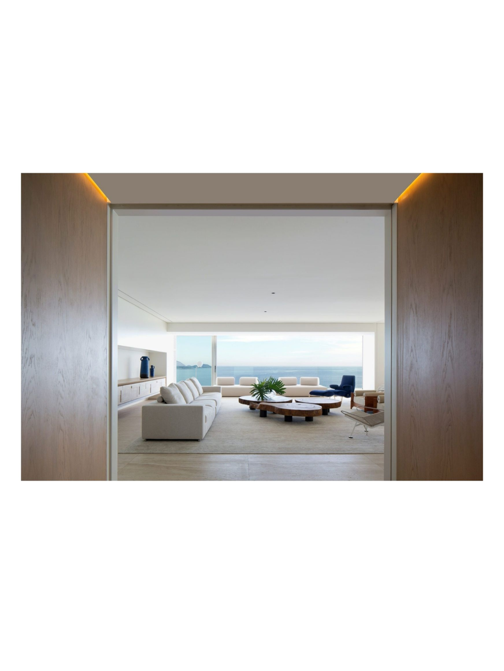 Book Arthur Casas: Architecture, featuring a modern, minimalist living room with an ocean view. The room showcases light-colored furniture, unique wooden coffee tables, and a dark blue accent chair, framed by light wood door jambs with warm backlighting. This architecture book is perfect for interior design enthusiasts and professionals seeking inspiration.