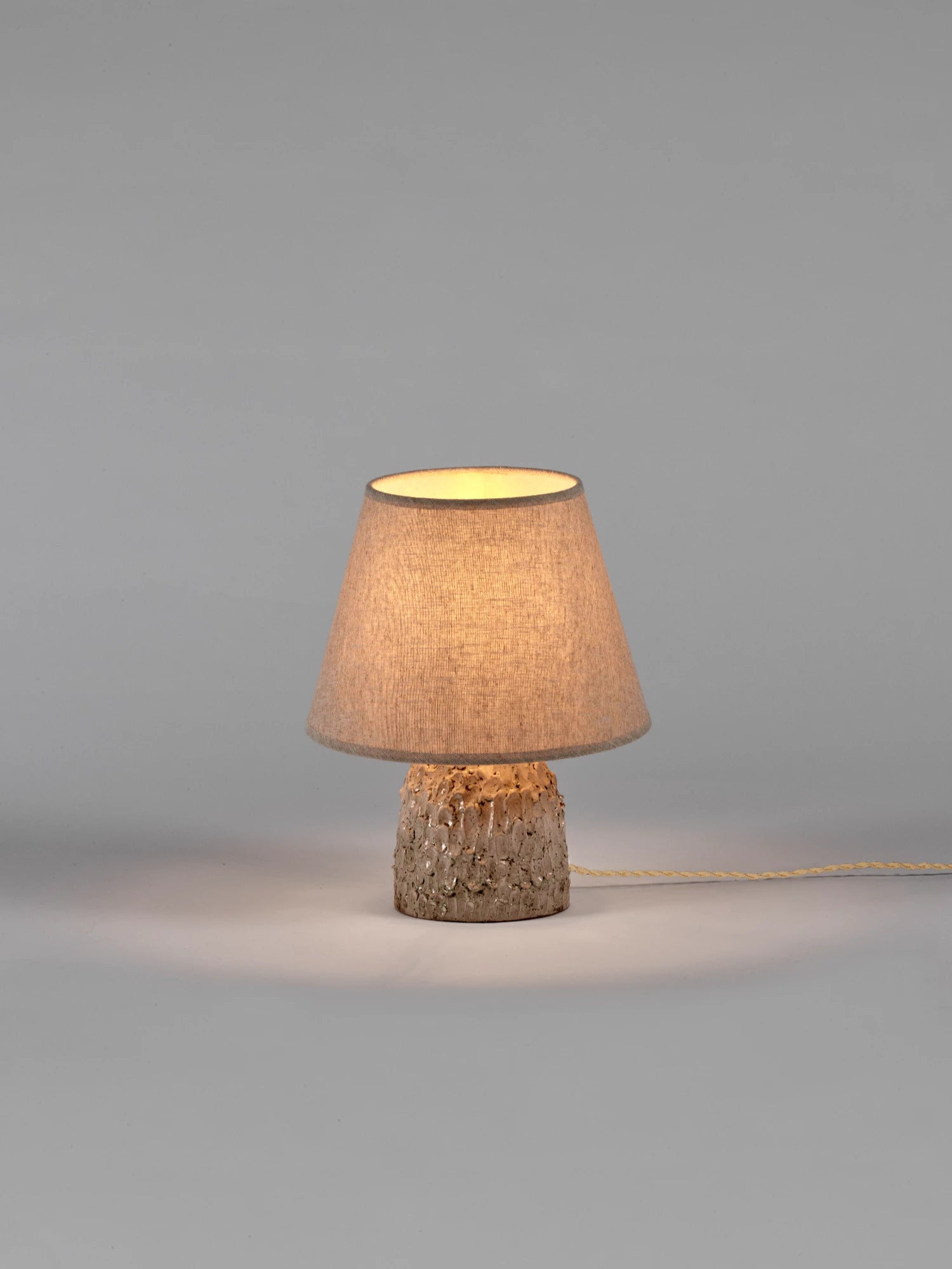 A small Tronco ceramic table lamp with a light beige/off-white, highly textured, glossy base and a conical, natural linen-colored lampshade. The light is switched on, giving the shade a warm glow. The lamp is photographed against a neutral gray background with a light-colored electrical cord extending to the right.